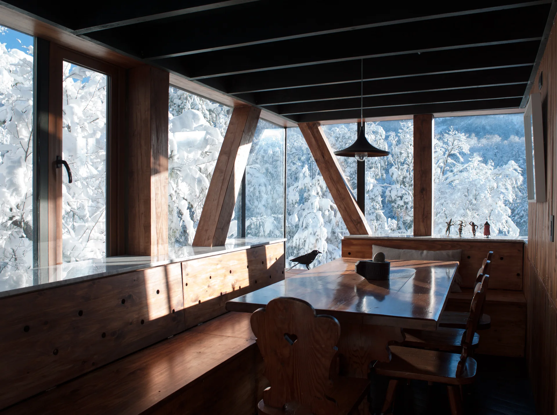 A rustic dining room features a wooden table and matching built-in benches set against large windows overlooking a snow-covered forest.