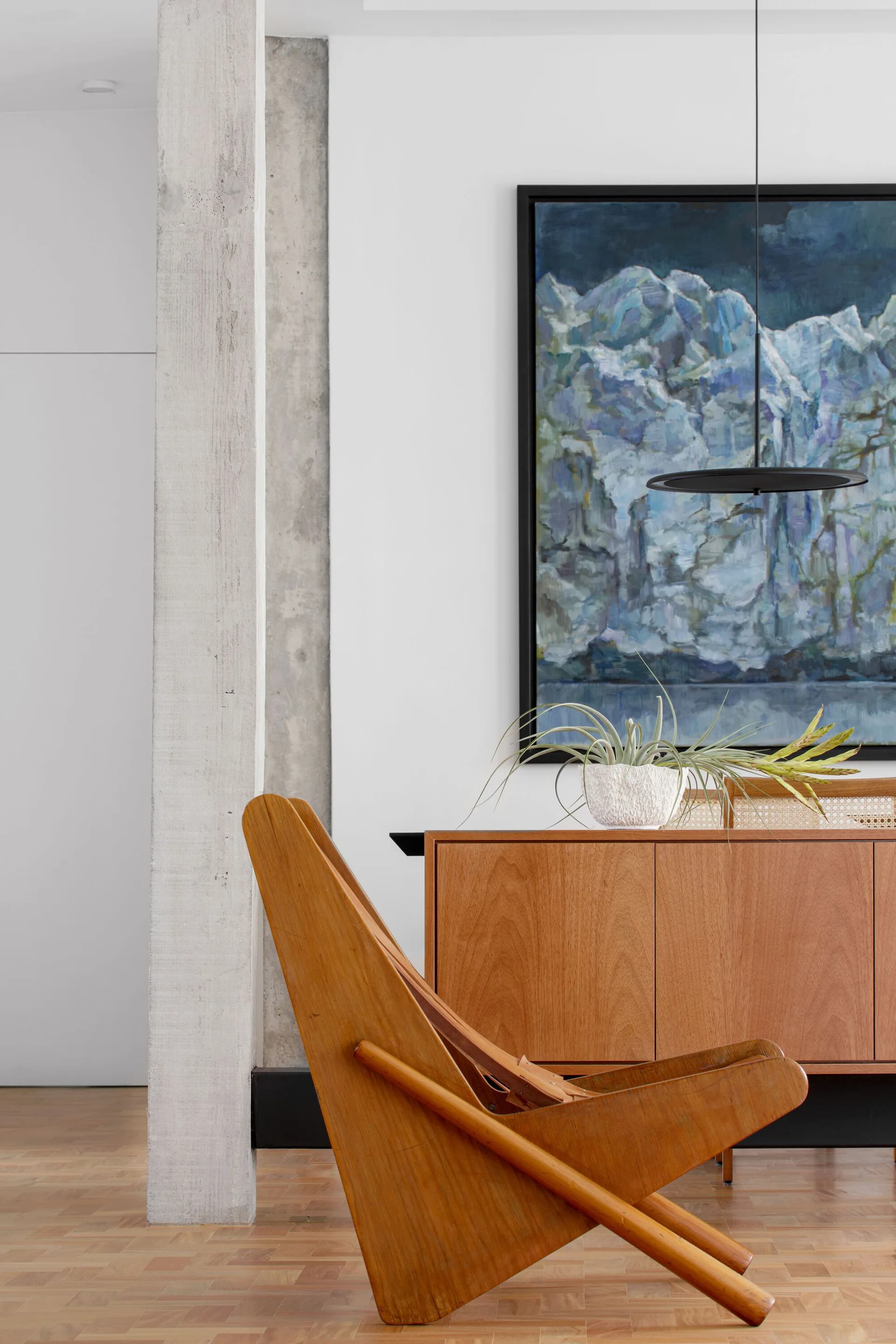 A sculptural mid-century wooden lounge chair sits near a wood sideboard with cane details under a large blue landscape painting next to a concrete column.