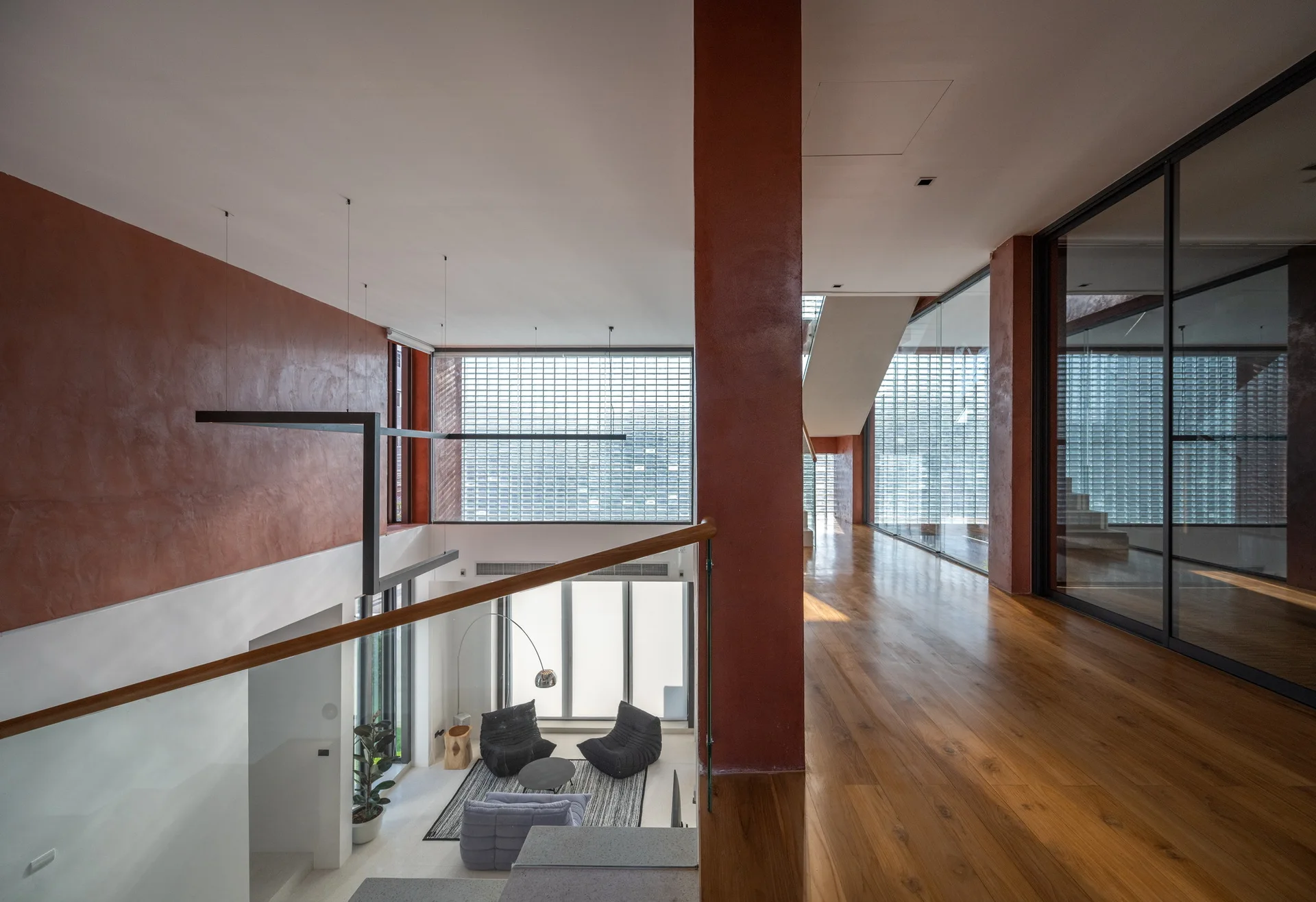 An upper-level wooden walkway looks down into a double-height living room featuring textured terracotta walls and a large glass block window.