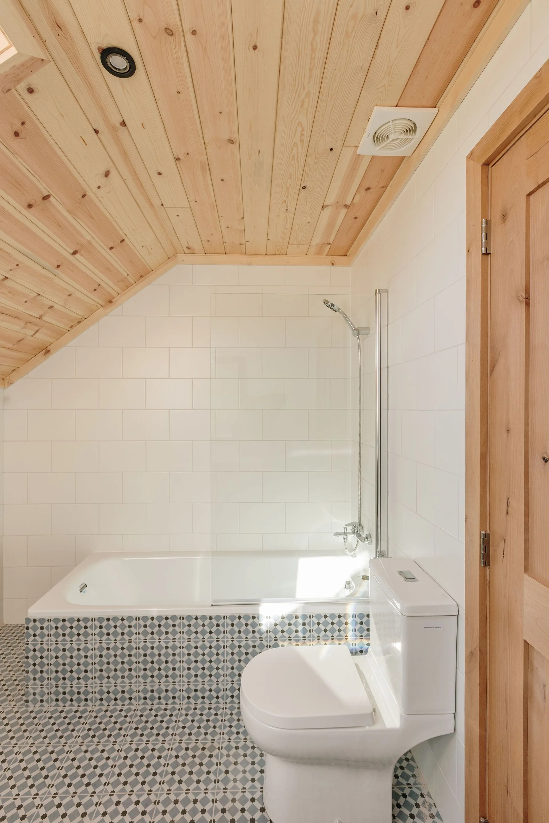 A compact bathroom with a sloped wood plank ceiling, white subway tiled walls, and geometric pattern tiles on the floor and bathtub apron.