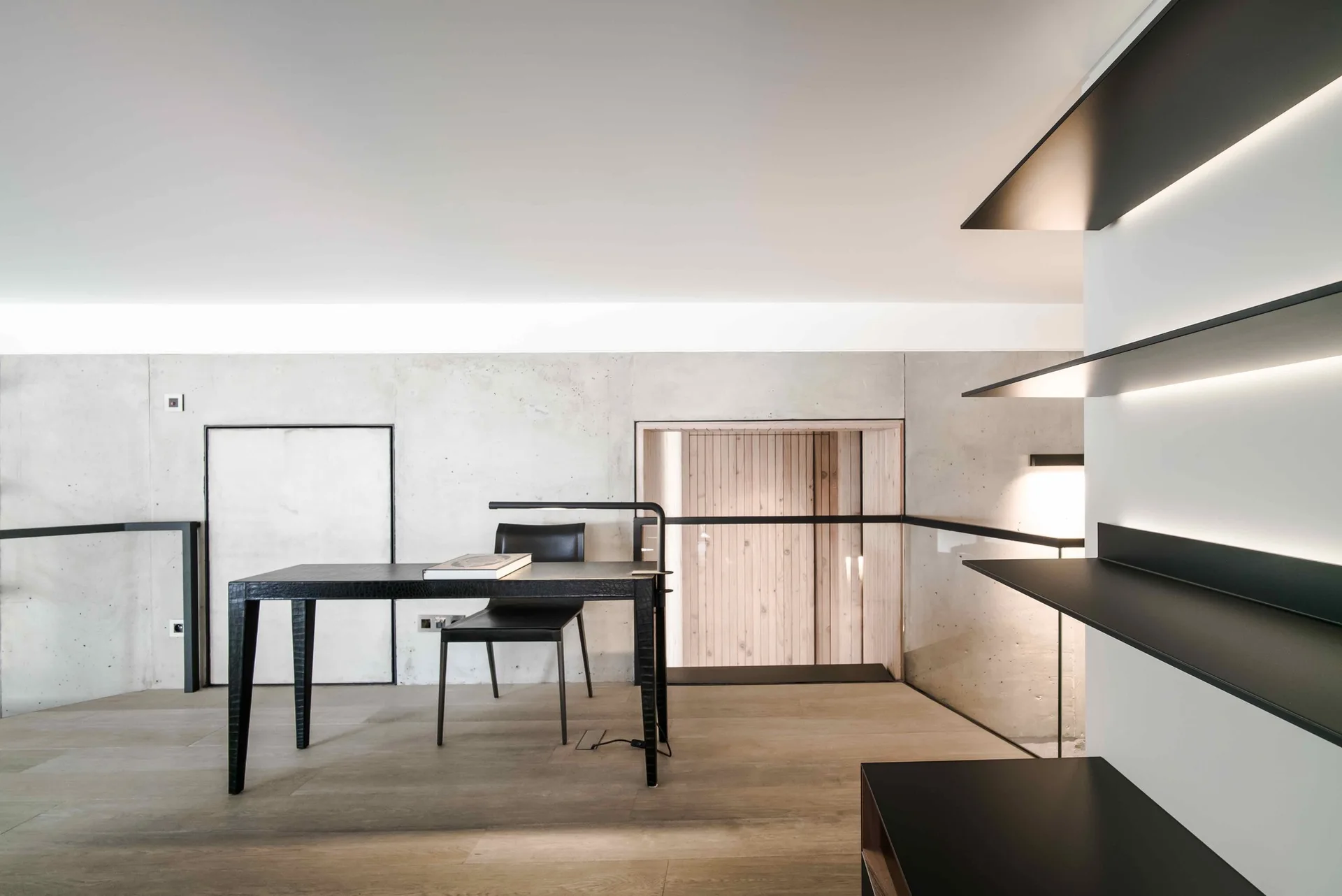 A minimalist home office features a black desk and chair set against a raw concrete wall next to floating black shelves with integrated light strips.