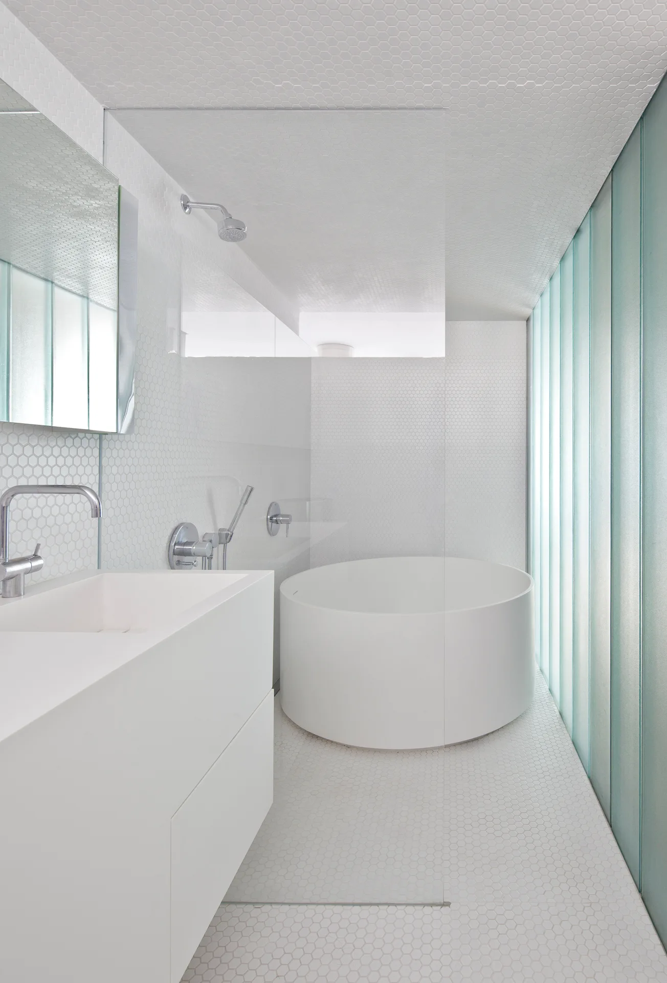 A minimalist white bathroom featuring a circular soaking tub, a floating white vanity, and small hexagon tiles covering the floor and walls.