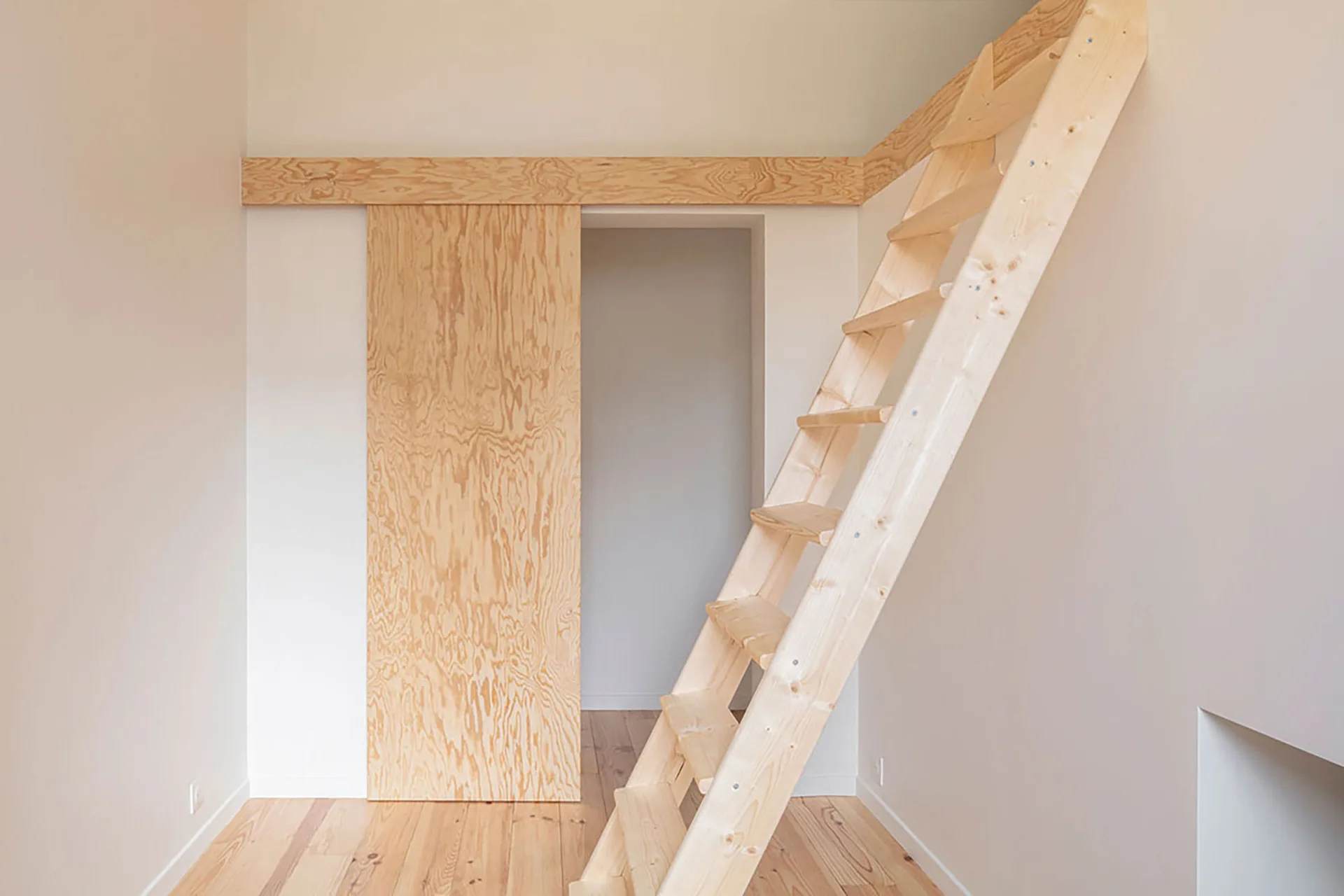 A minimalist white room featuring a light pine ladder staircase and a sliding plywood door with distinct wood grain patterns.