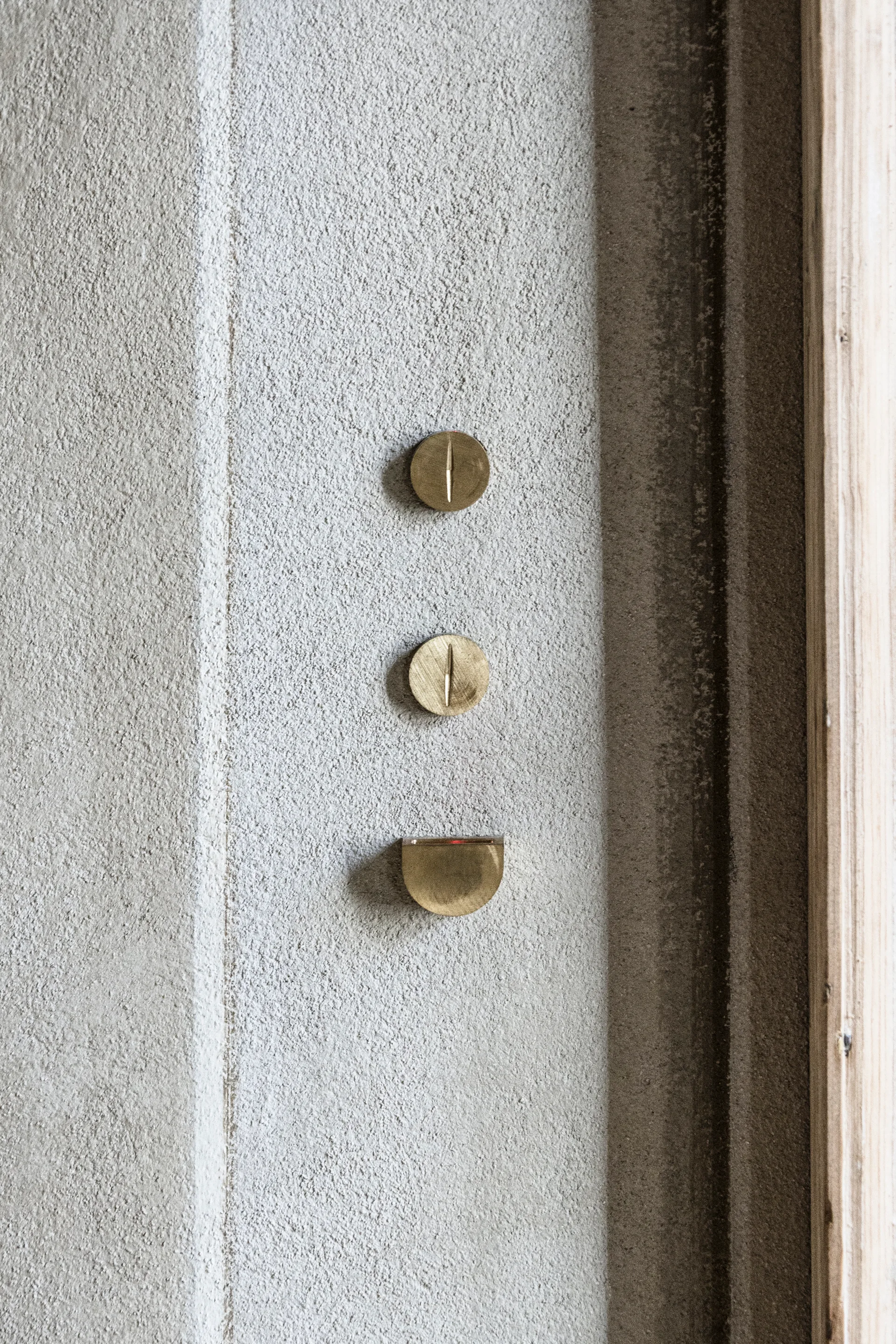 Close-up of three minimal brass switches and buttons mounted on a textured beige plaster wall next to a light wood door frame.