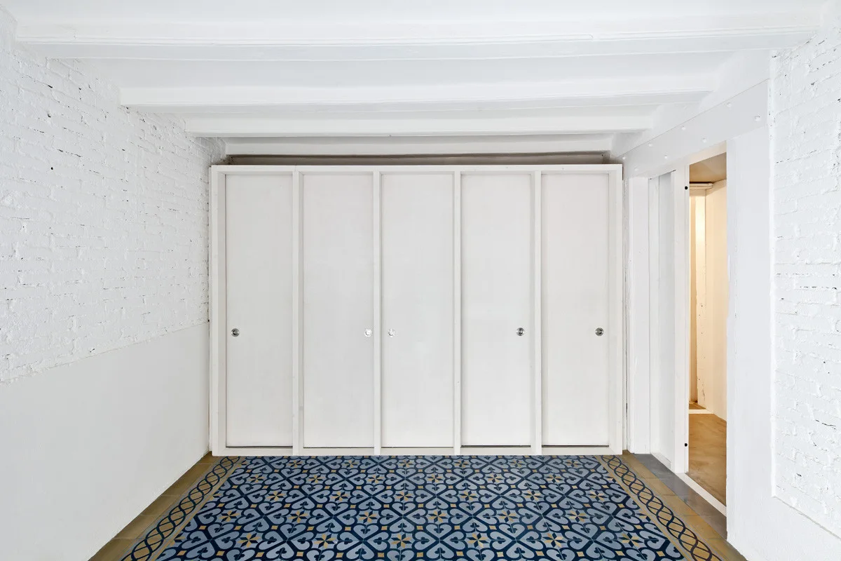 Minimalist room featuring white floor-to-ceiling built-in cabinets, painted brick walls, exposed ceiling beams, and a blue and yellow patterned tile floor.