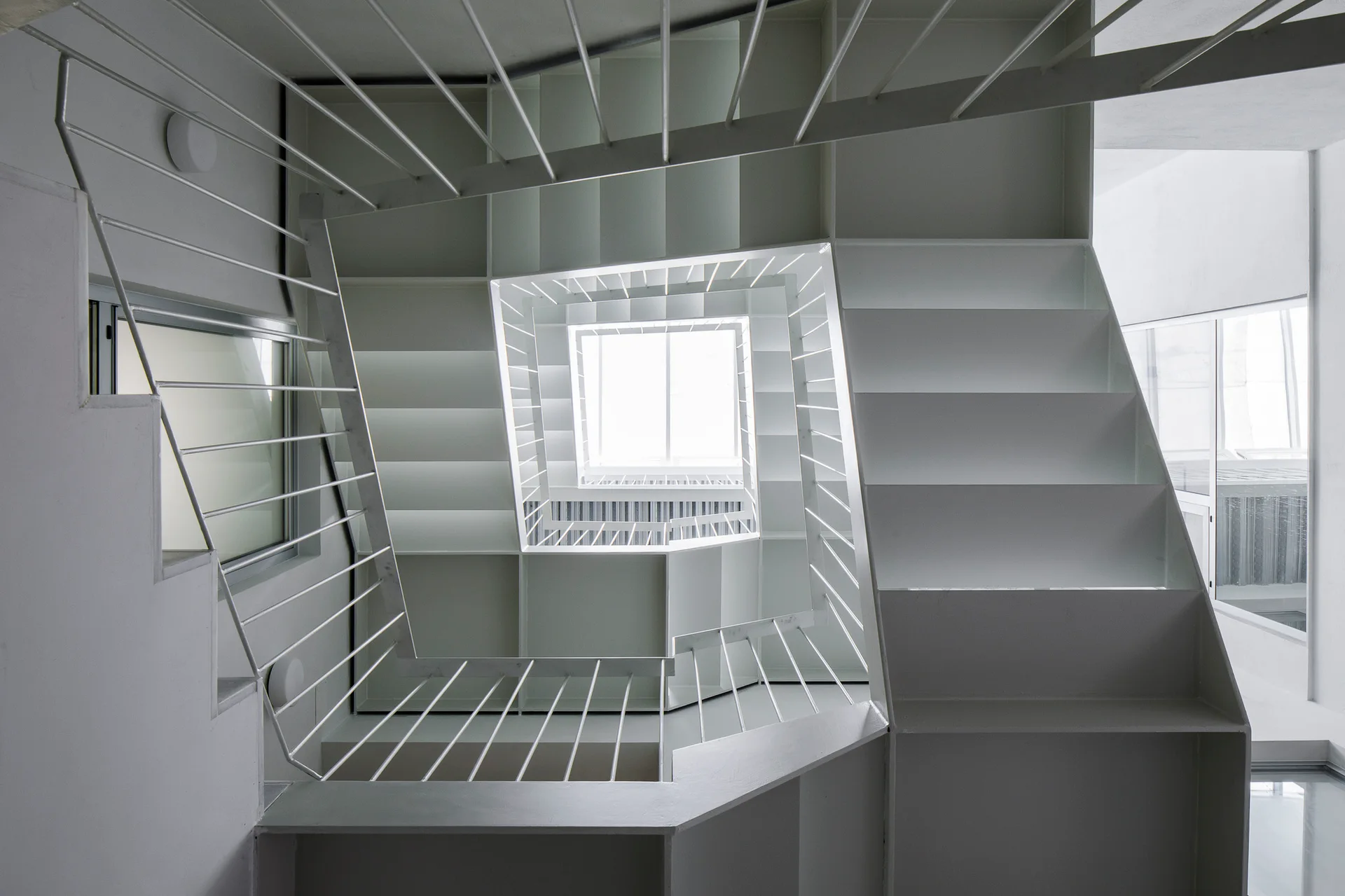 A modern minimalist white metal staircase spiraling around a square central void with thin metal rod railings and natural light.