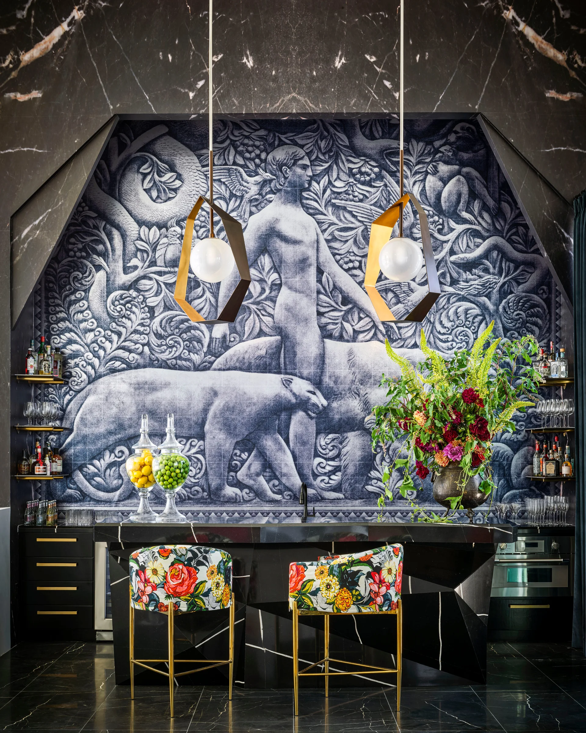 A home bar features a black marble counter, two colorful floral bar stools, a large monochrome mural, and gold pendant lights.