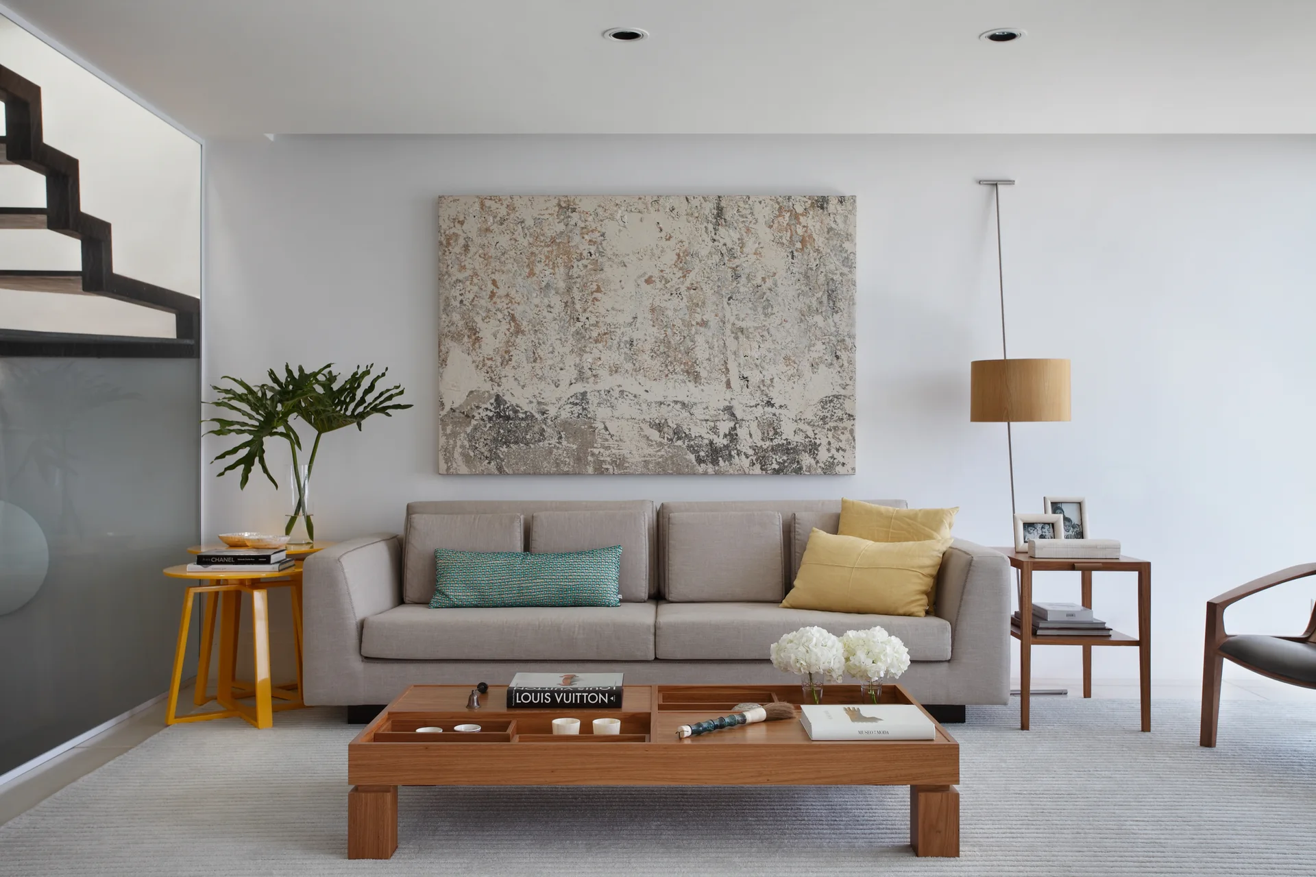 A light grey sofa sits in a neutral living room between a yellow accent table and a wooden side table, with a compartmentalized coffee table in front.