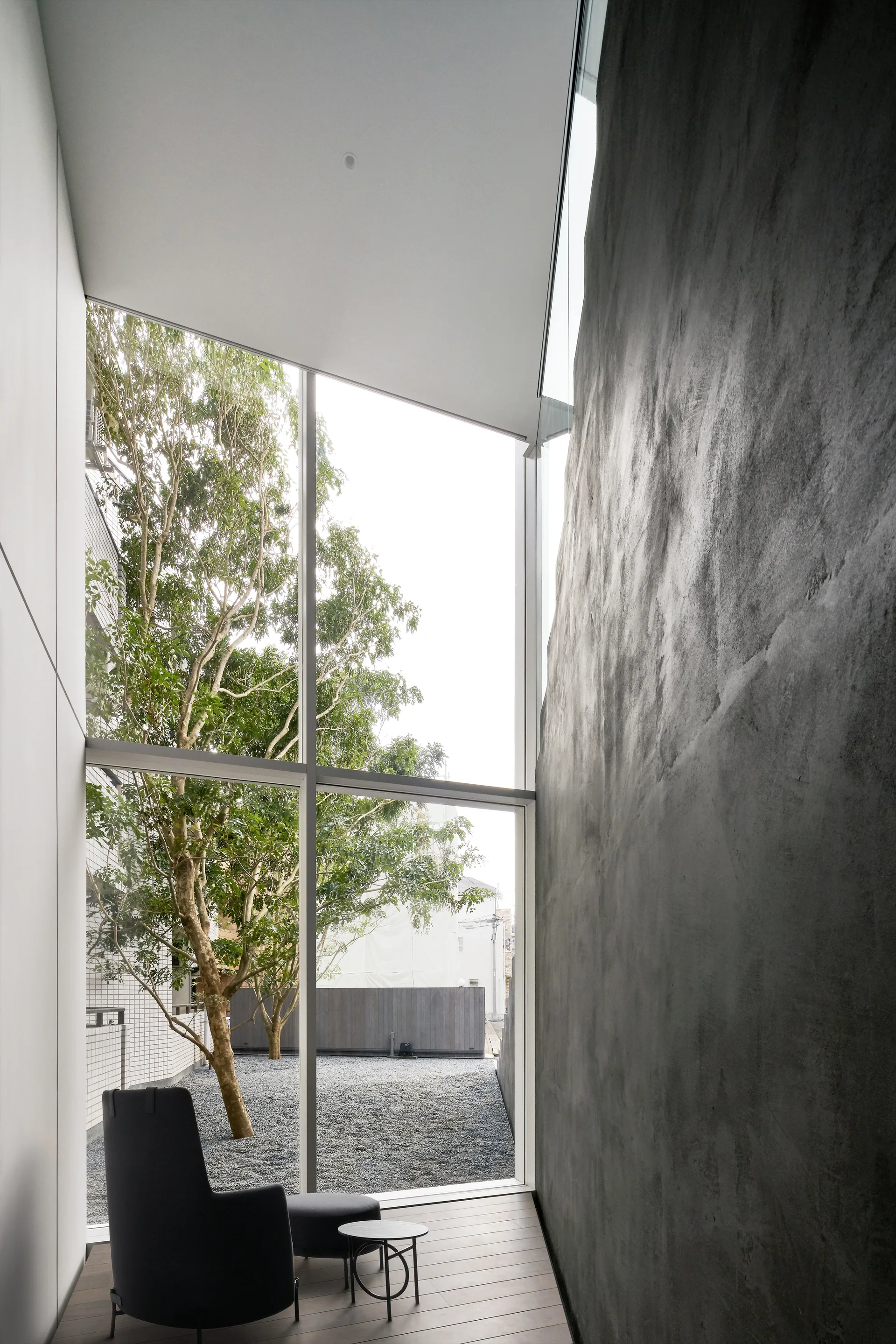A minimalist nook features a dark lounge chair next to a tall, textured concrete wall and a window framing a gravel courtyard.