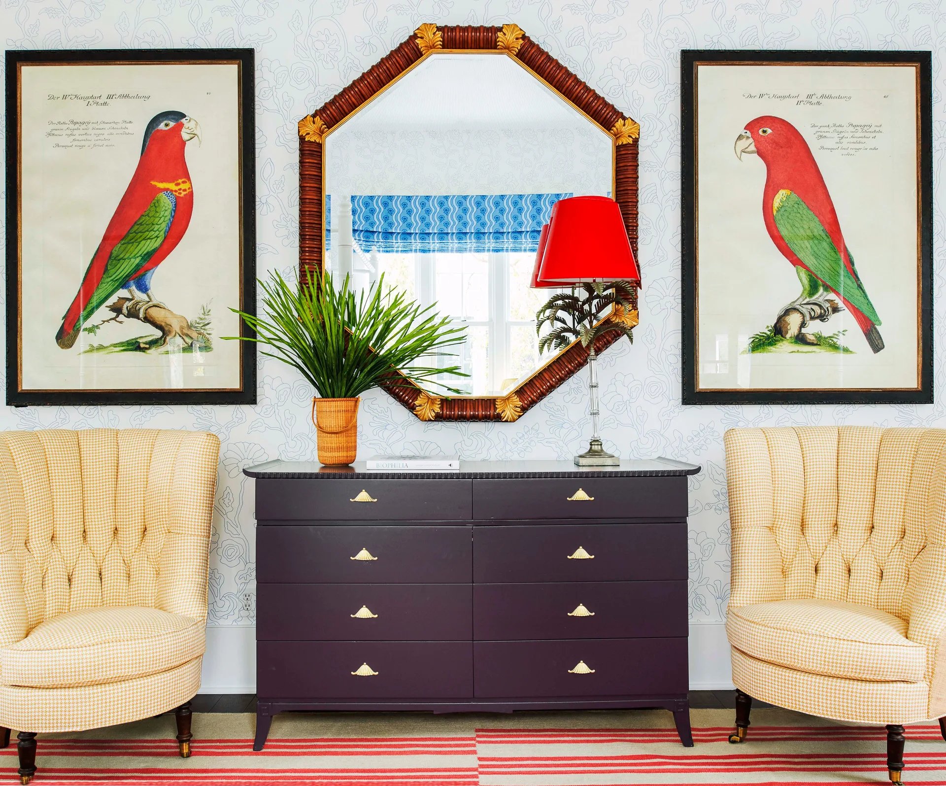 A dark purple dresser is flanked by yellow houndstooth armchairs under an octagonal mirror and two framed botanical parrot prints.
