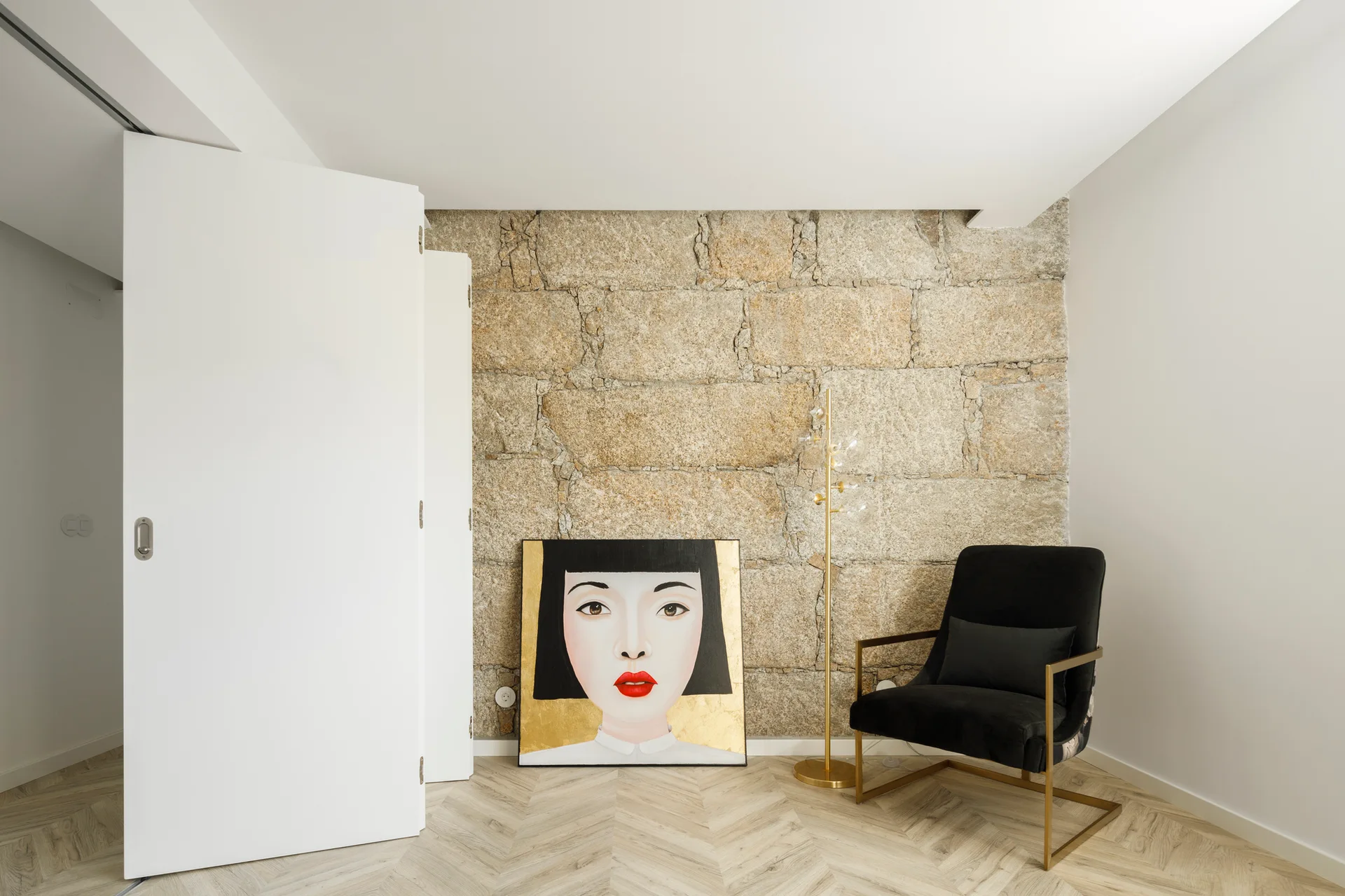 A living room corner features an exposed stone block wall, chevron wood flooring, a black armchair, and a large portrait painting leaning on the floor.