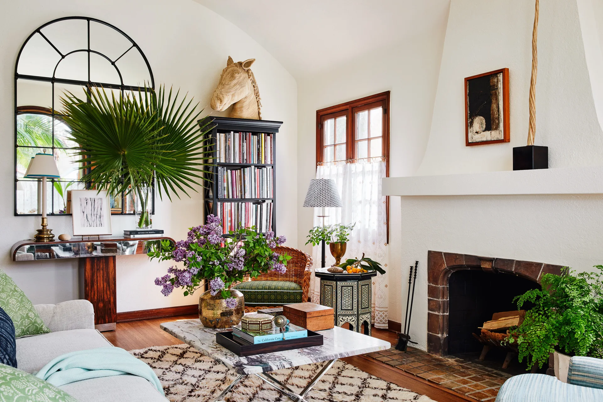 An eclectic living room features a white plaster fireplace, an arched wall mirror, a tall black bookshelf, and a marble coffee table with a lilac bouquet.