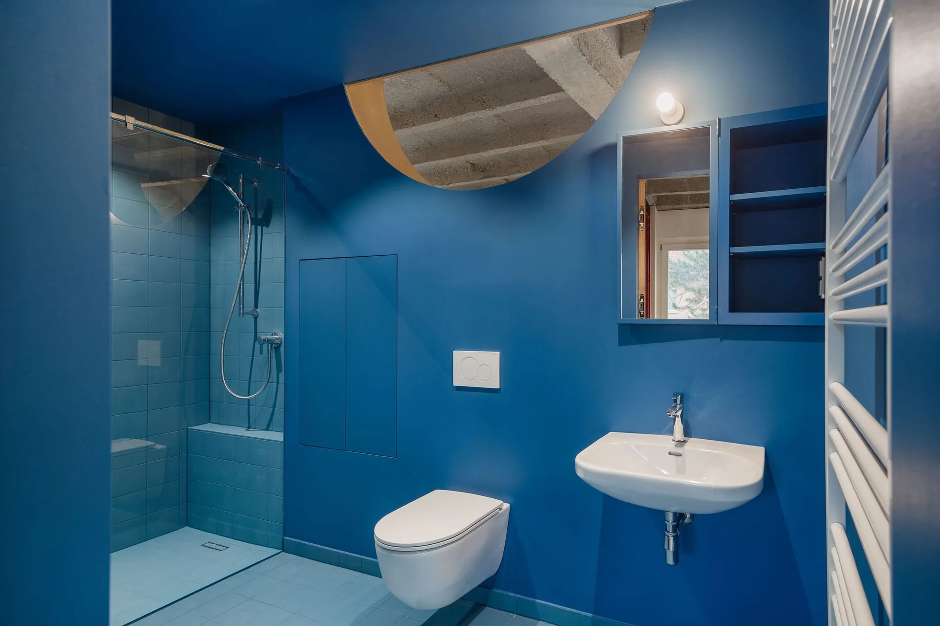 A modern bathroom featuring blue monochromatic walls and tiled floors with a wall-hung toilet, white sink, and a curved ceiling cutout revealing concrete.