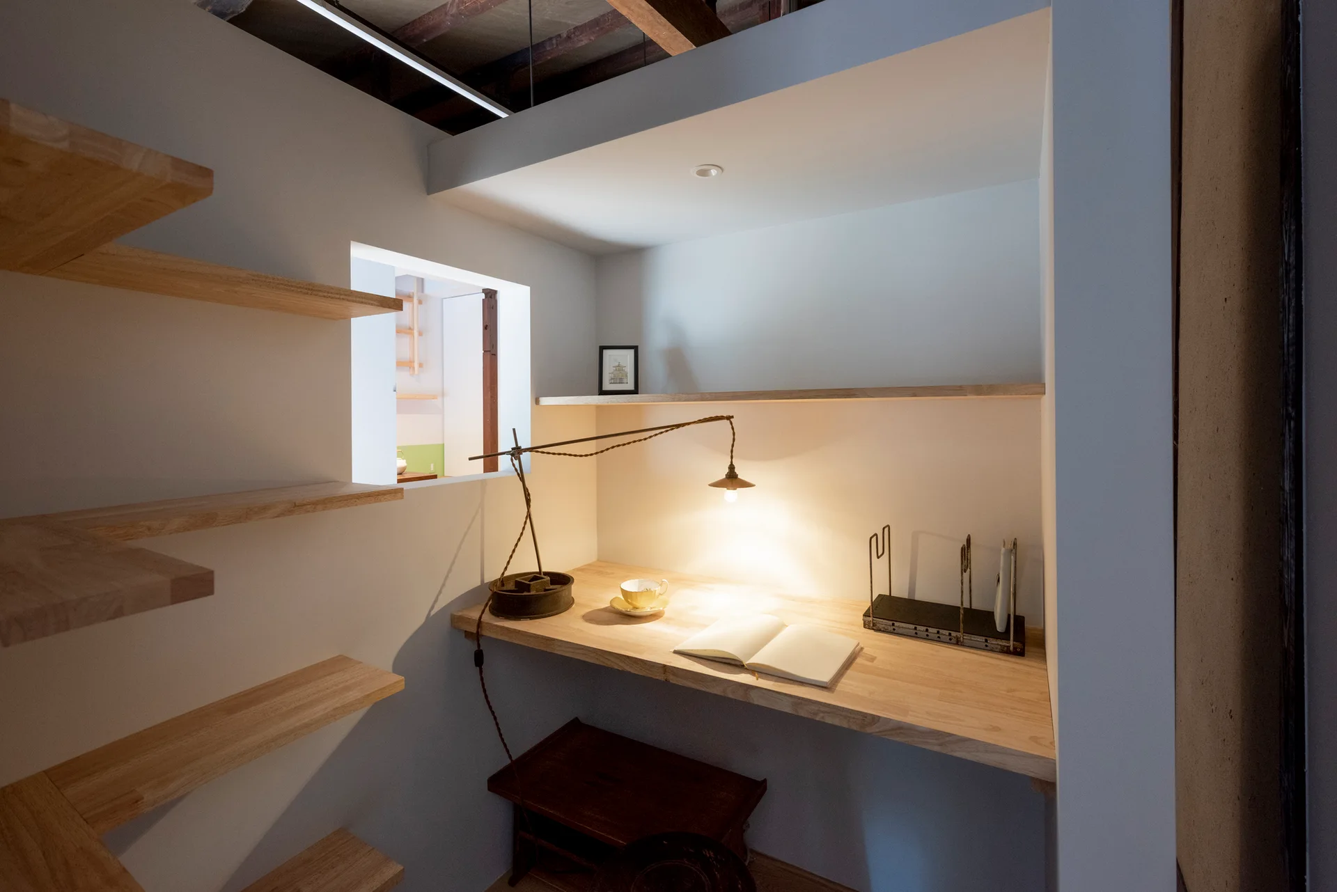 A compact home office alcove with a light wood floating desk, matching open shelving, and a small square cutout window.