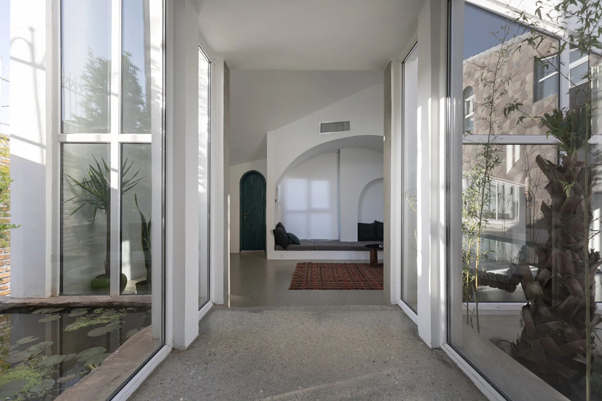 A concrete walkway flanked by large glass windows looks out onto courtyards with a pond, leading into an indoor room with a built-in daybed.