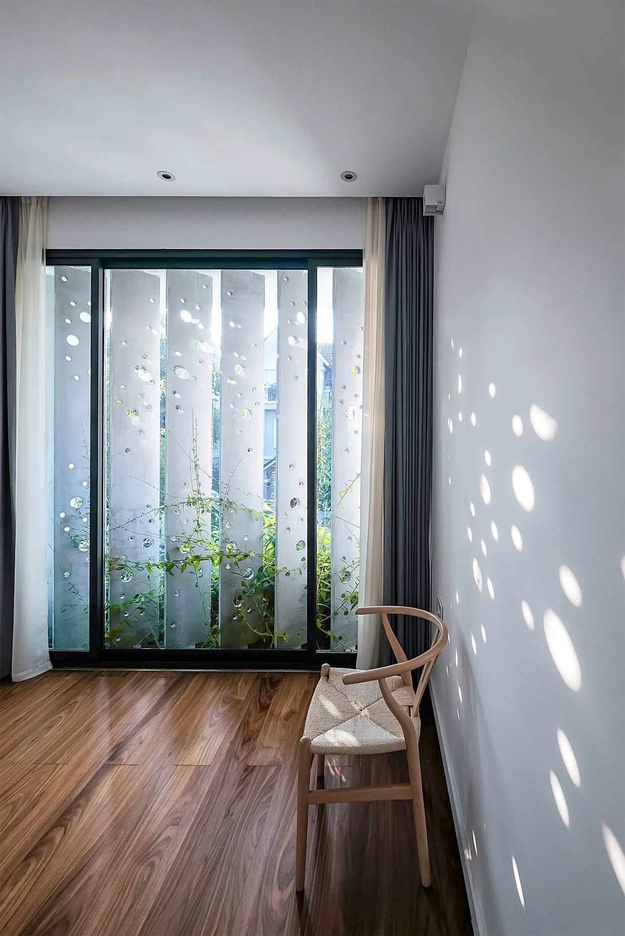A wooden chair sits on a hardwood floor near a window with a perforated screen casting circular light patterns on a white wall.