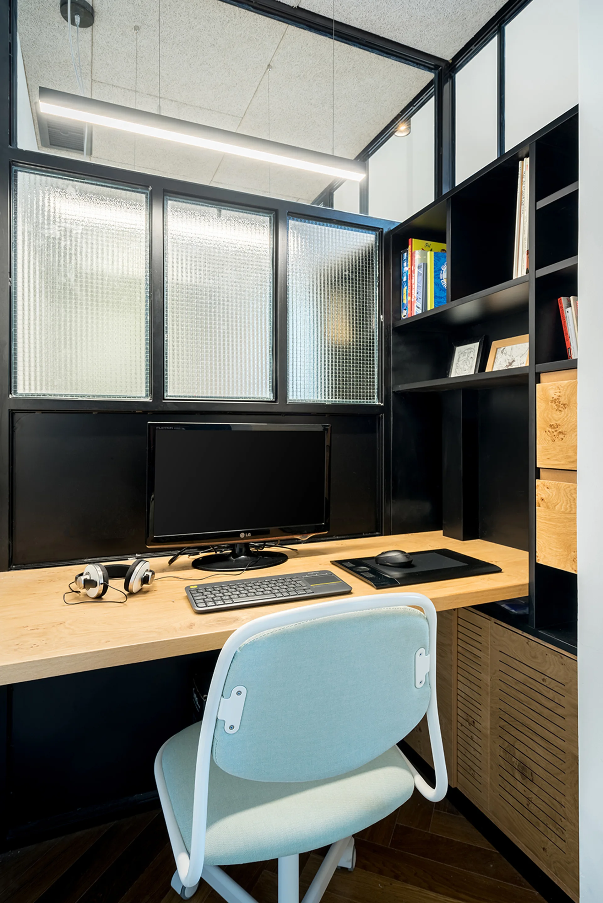 A compact home office features a wood desk, a light blue chair, black shelving, and a black-framed textured glass partition wall.