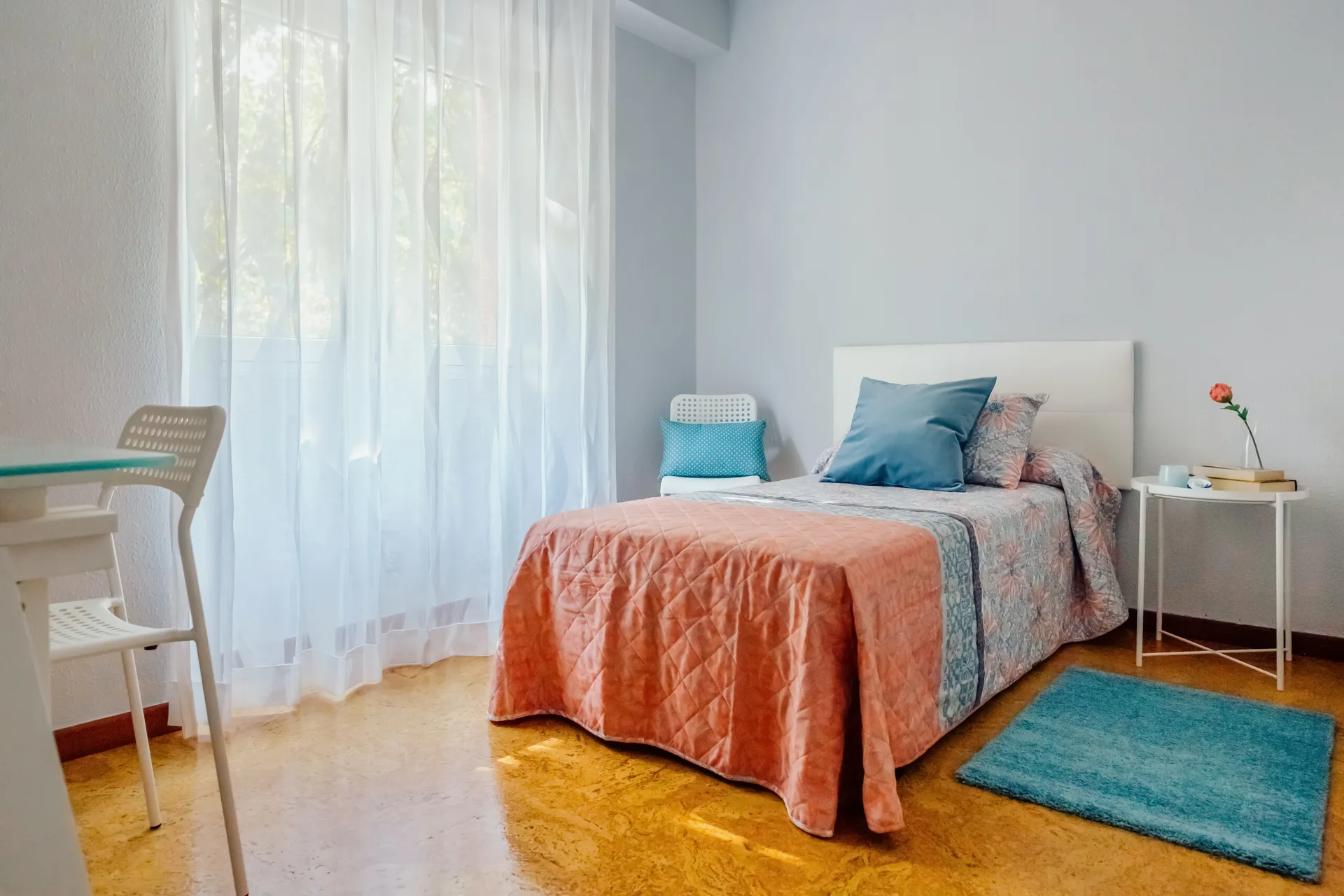 A single bed with coral quilted bedding and a blue pillow is set against light blue walls near a window with sheer white curtains.