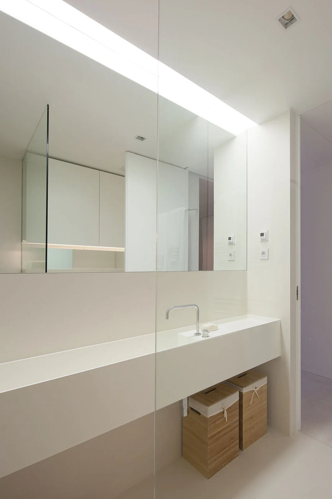 A minimalist white bathroom featuring a floating trough sink, a large rectangular mirror, and two wooden laundry baskets underneath.