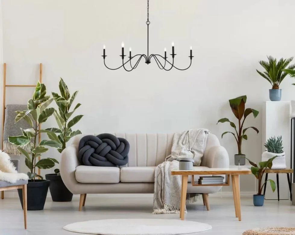 A light beige sofa with a grey knot pillow is flanked by potted houseplants beneath a black metal candle chandelier in a bright minimalist living room.