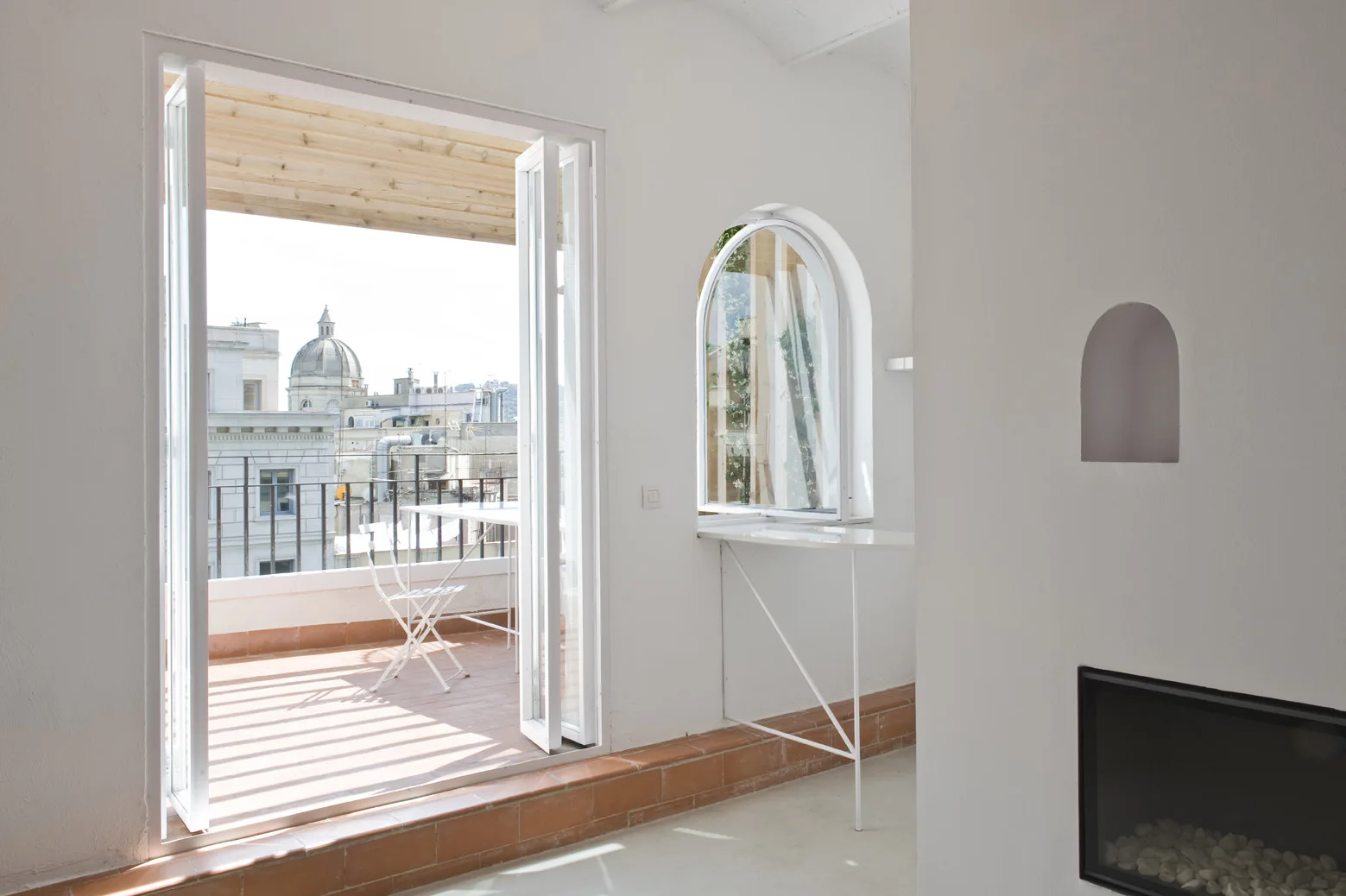 A bright white minimalist room features an arched window and open folding doors leading to a sunny terracotta terrace with a bistro table.