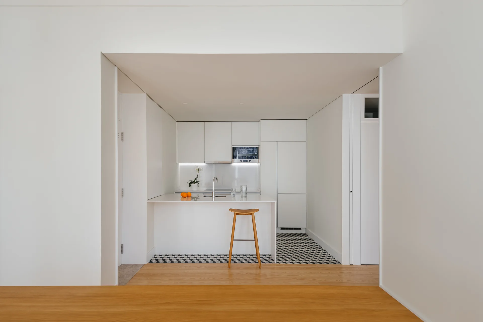 A minimalist white kitchen alcove features a white island with a single wooden stool, set against geometric patterned floor tiles.