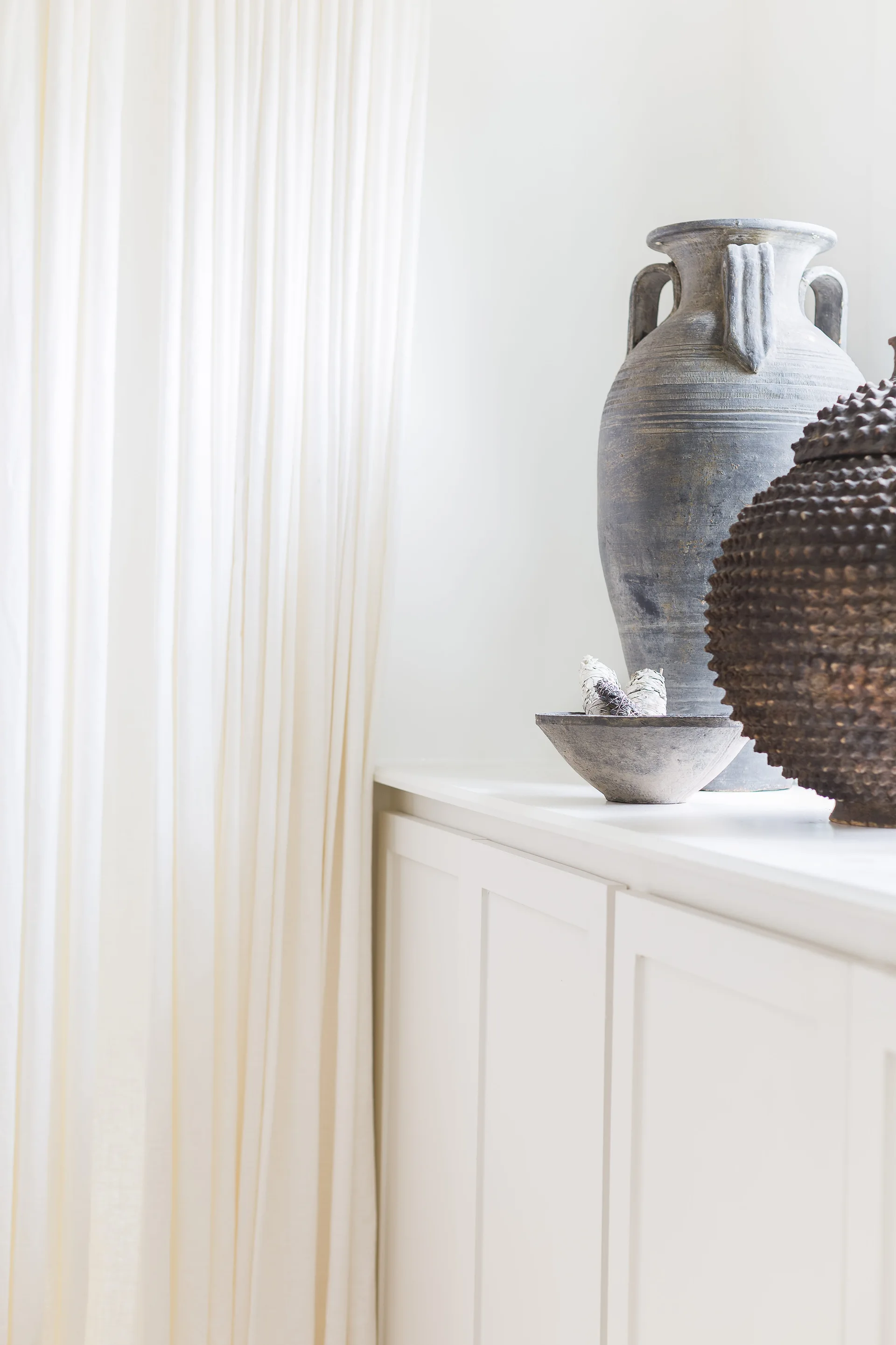 A close-up of a white cabinet topped with a rustic grey stone urn, a small bowl, and a dark textured orb next to soft white curtains.