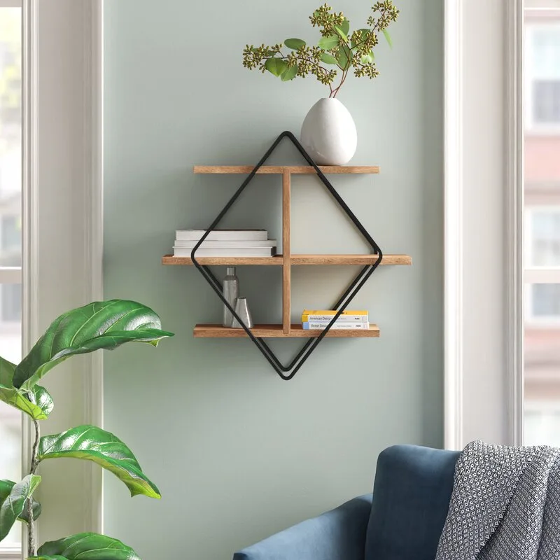 A diamond-shaped black metal and wood wall shelf displays a white vase and books against a light sage green painted wall.