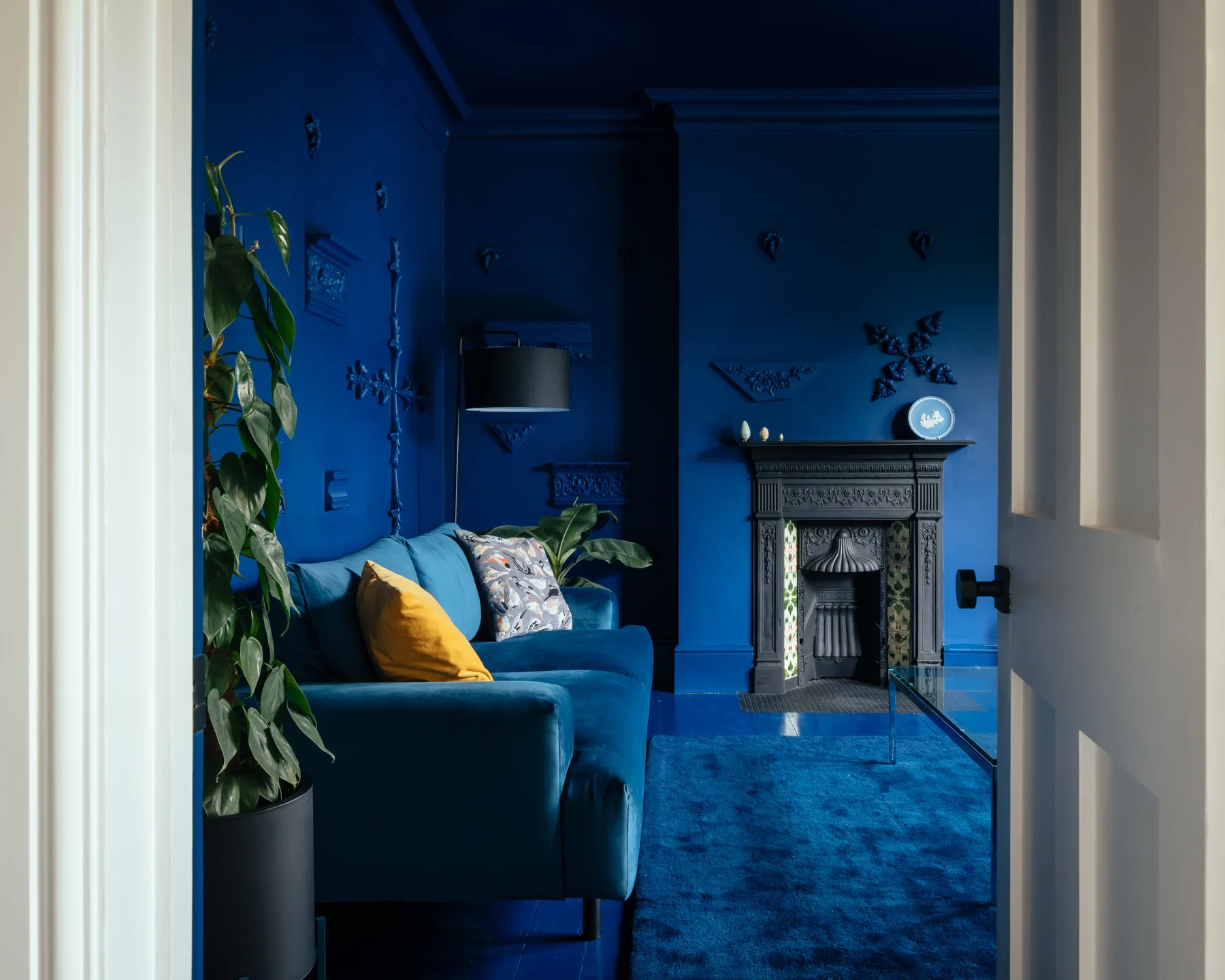 A monochromatic deep blue living room features a matching velvet sofa, a black ornate fireplace, and wall moldings seen through a white doorway.