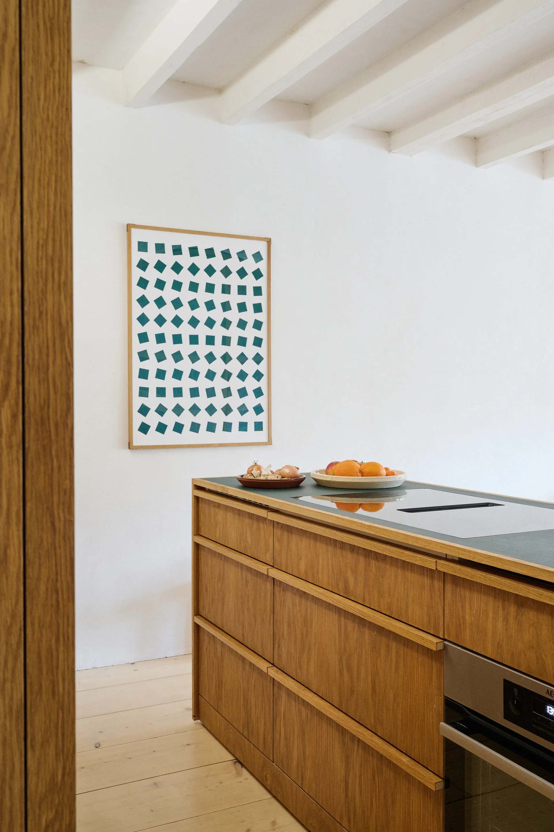 A natural wood kitchen island with a green countertop stands in a white room with exposed ceiling beams and geometric wall art.