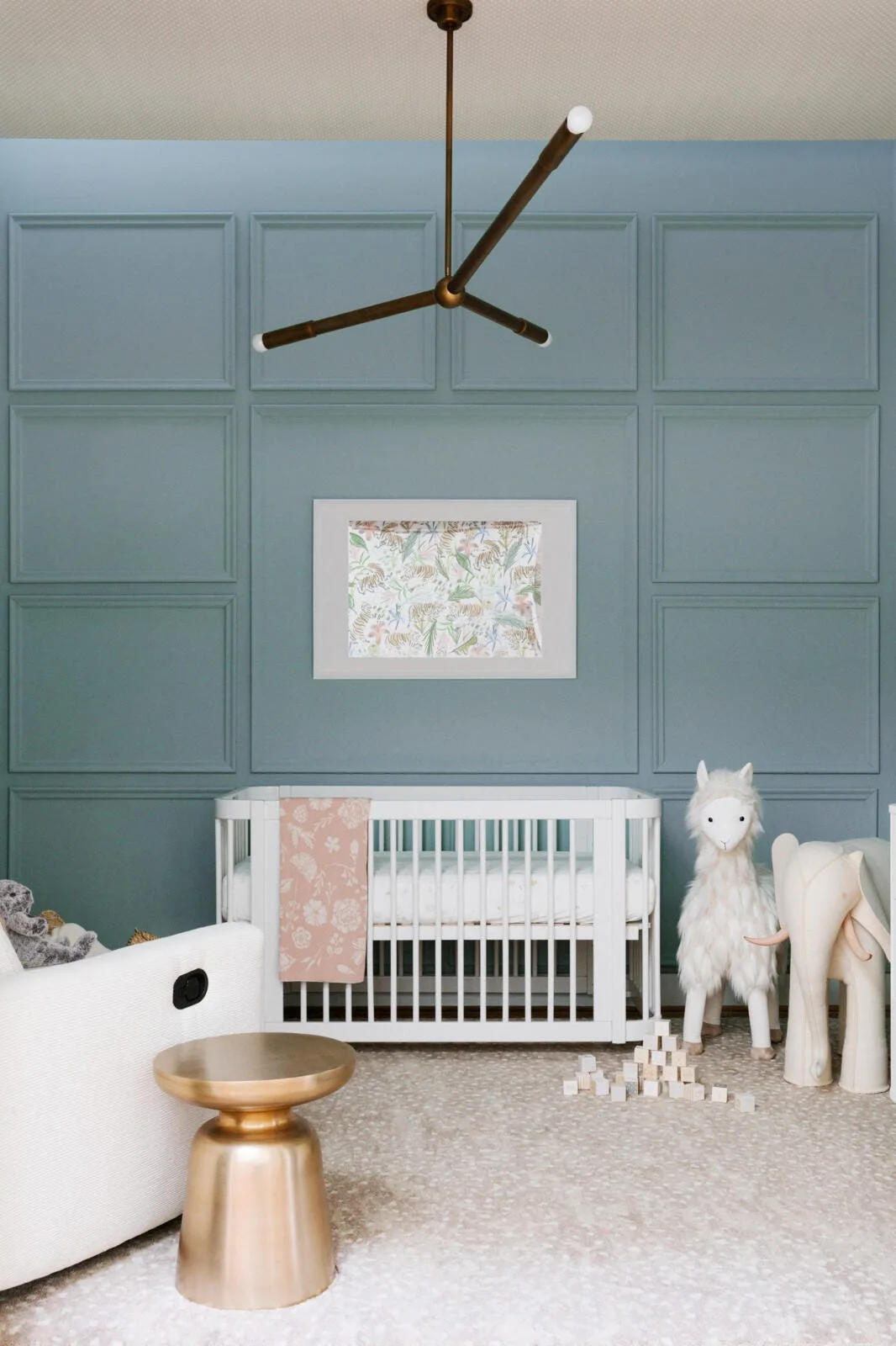 A baby nursery features a white wooden crib placed against a dusty blue grid-paneled wall, accented by a gold side table and plush animals.
