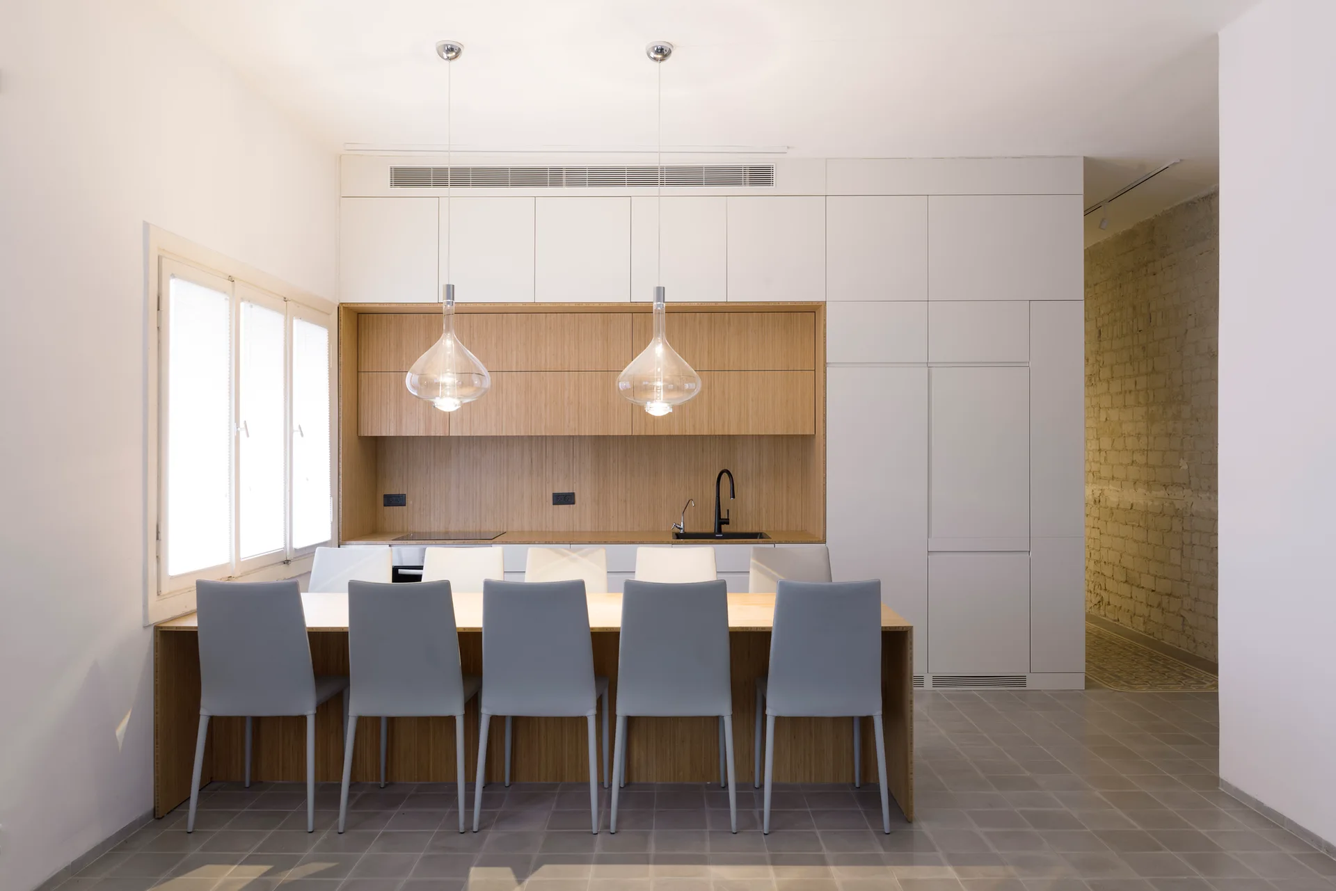 A minimalist kitchen features white slab cabinetry, a recessed wooden niche, a wooden dining table with grey chairs, and glass pendant lights.