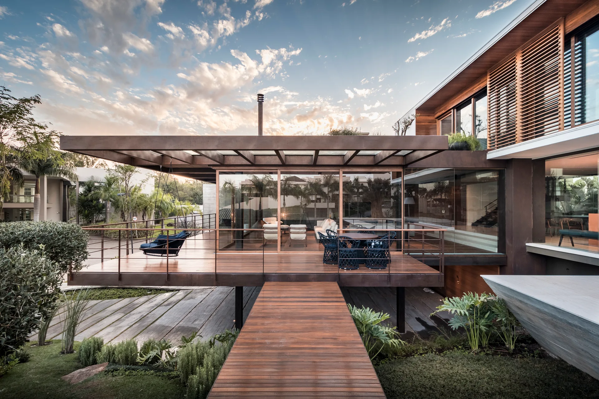 An elevated wooden deck with a matching walkway leading to a modern home featuring floor-to-ceiling glass walls and outdoor seating at dusk.