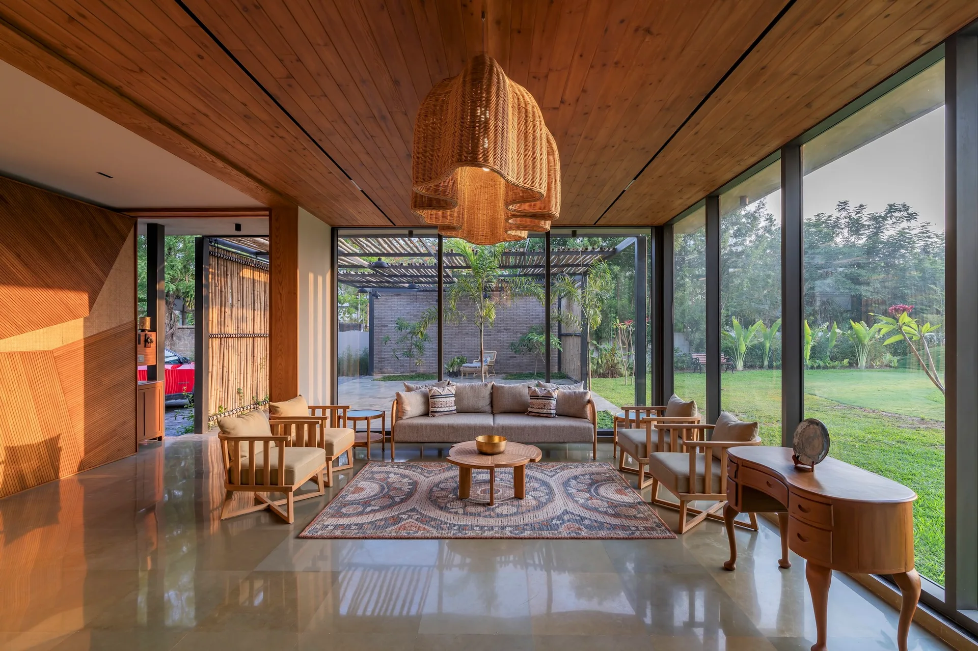 A living room featuring a wood plank ceiling, a large woven pendant light, wood-framed armchairs, and floor-to-ceiling glass walls showing a garden.