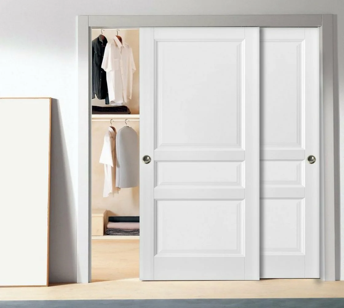 Two matte white sliding closet doors with recessed panels are partially open to reveal hanging clothes inside.