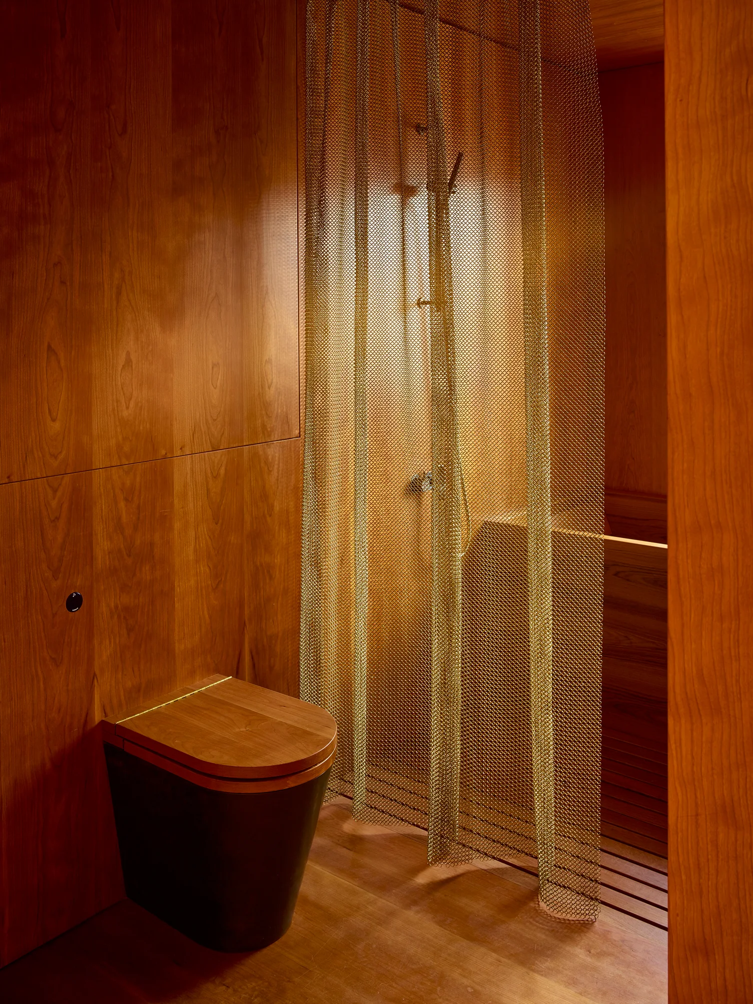 A minimalist bathroom features wood paneled walls, a wood-topped toilet, and a brass chainmail mesh shower curtain.
