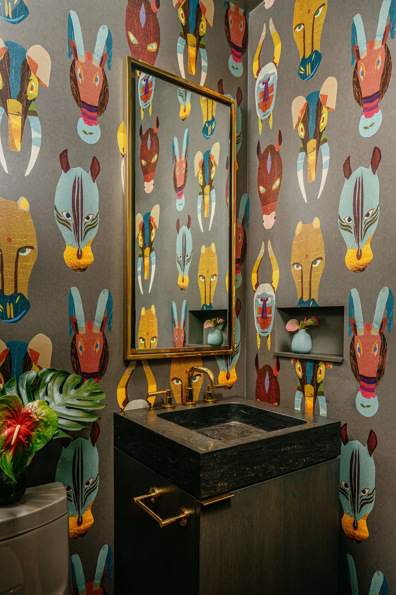 A compact powder room with colorful animal mask wallpaper, a dark stone vanity, a brass faucet, and a brass-framed mirror.