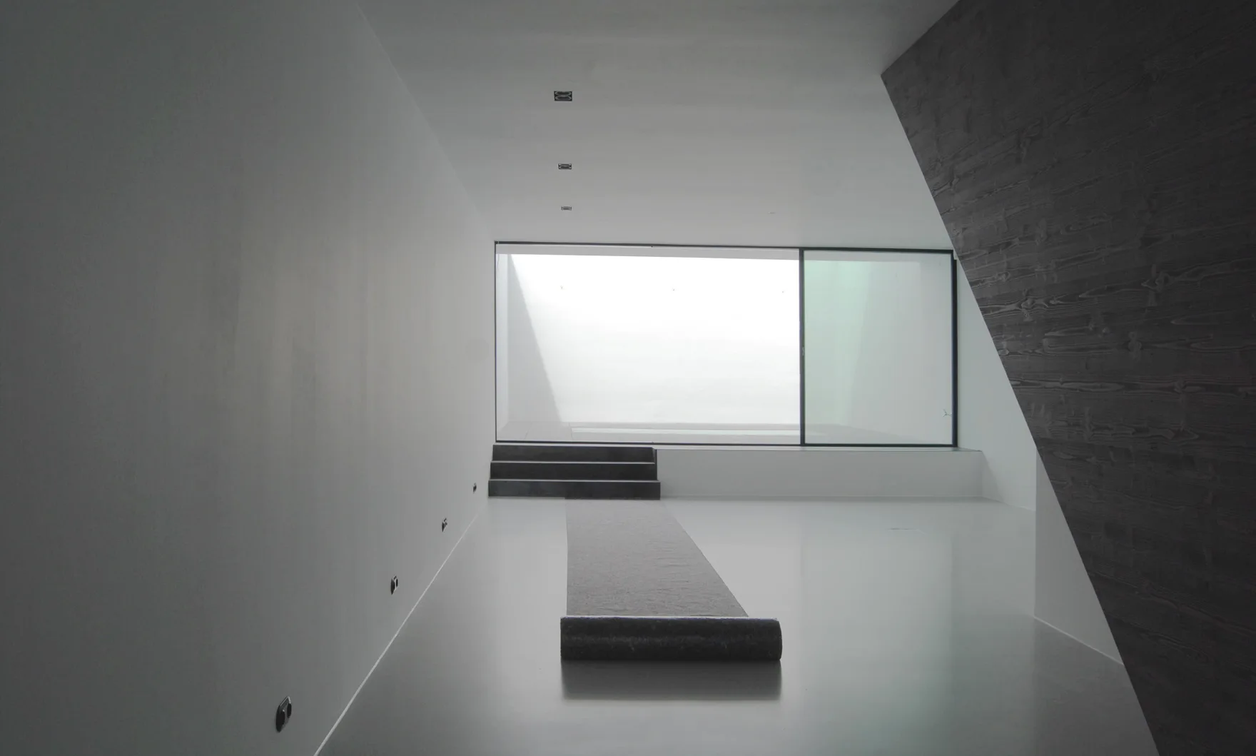 A minimalist white room featuring a slanted dark wood-grain wall, a grey floor runner, and steps leading to a large sliding glass window.