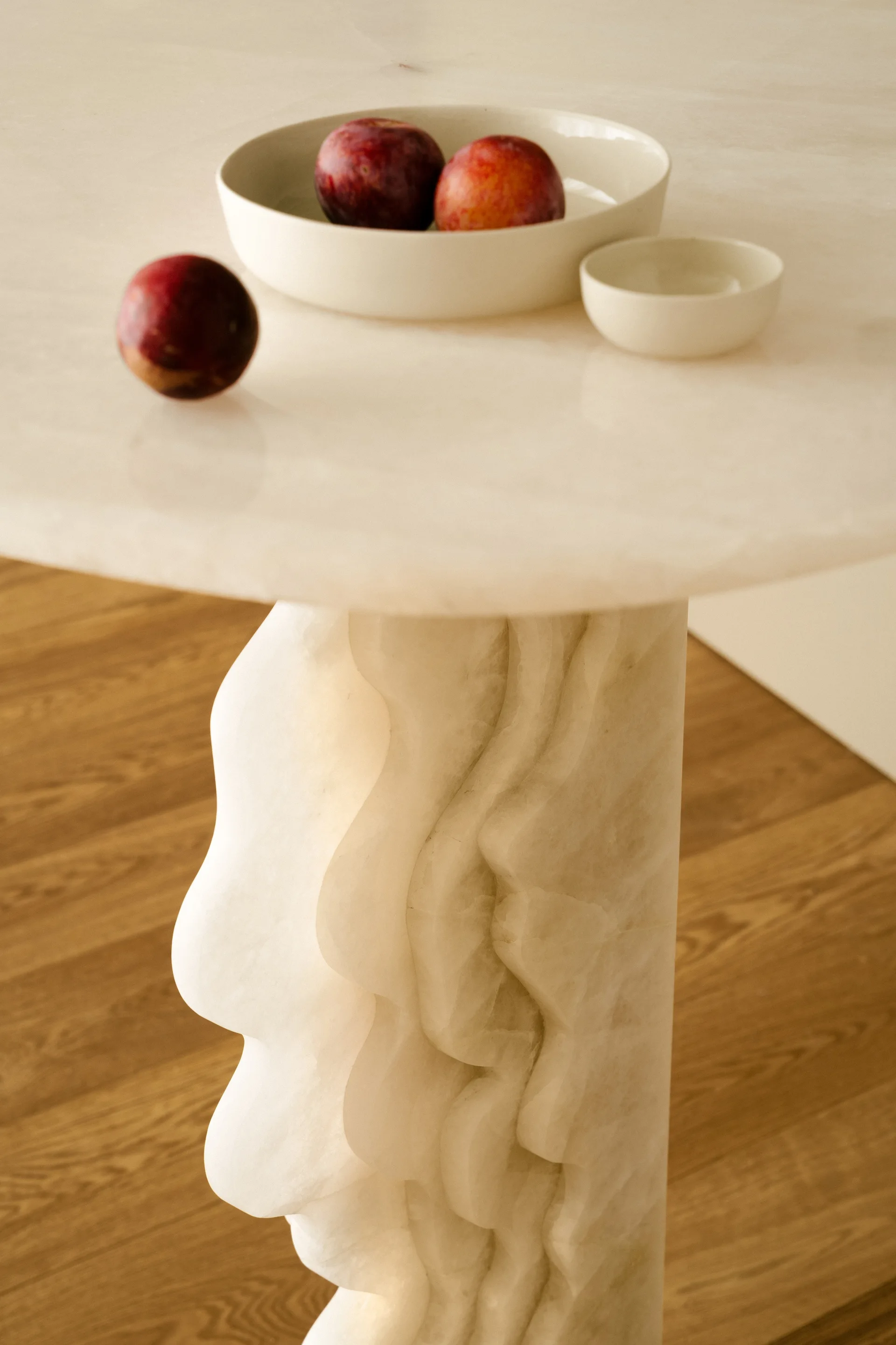 A close-up shot of a round stone table featuring a wavy sculptural pedestal base, with cream-colored ceramic bowls and red plums on its polished top.