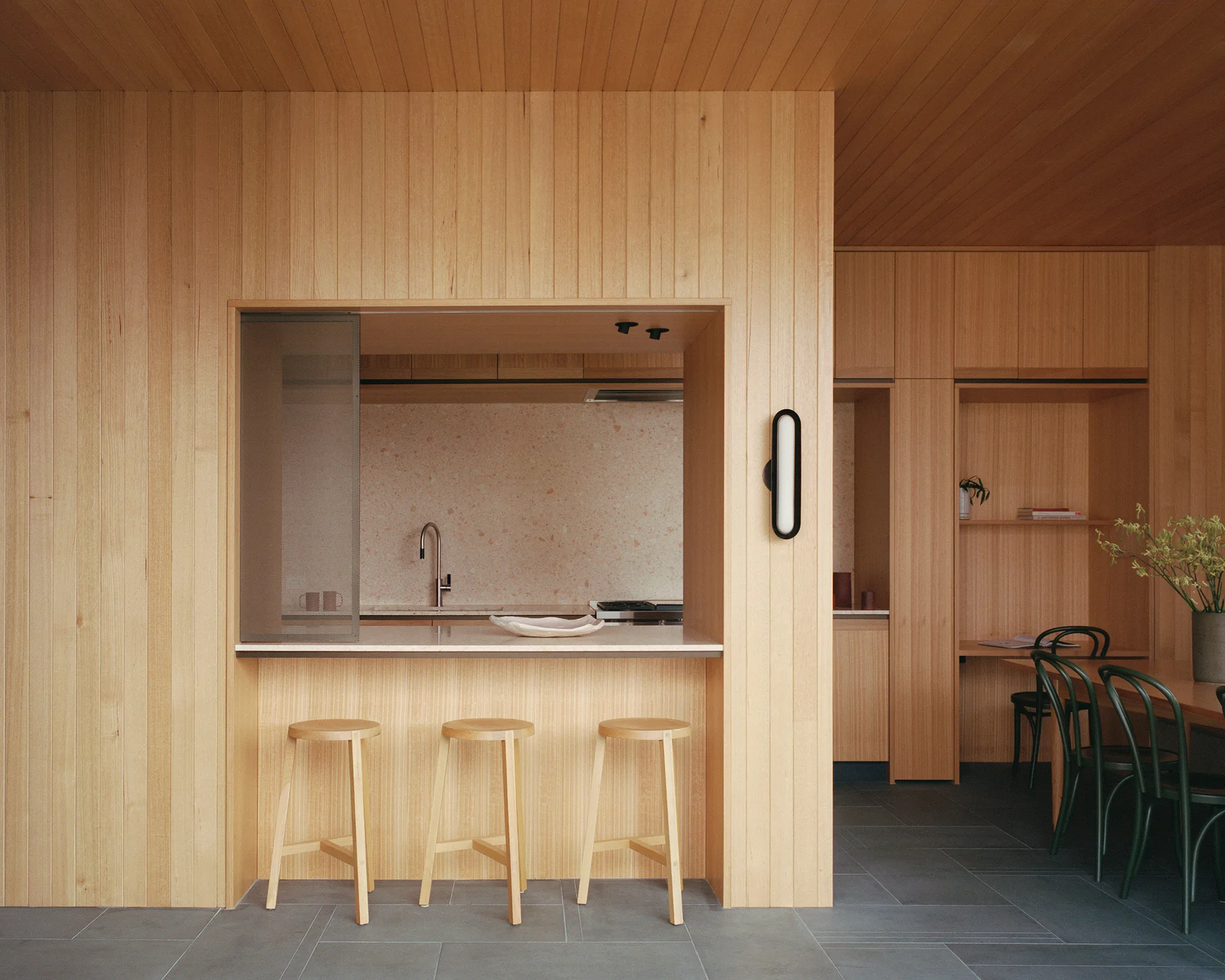 A minimalist kitchen features vertical light wood paneling, three wooden stools at a pass-through counter, and a speckled terrazzo backsplash.