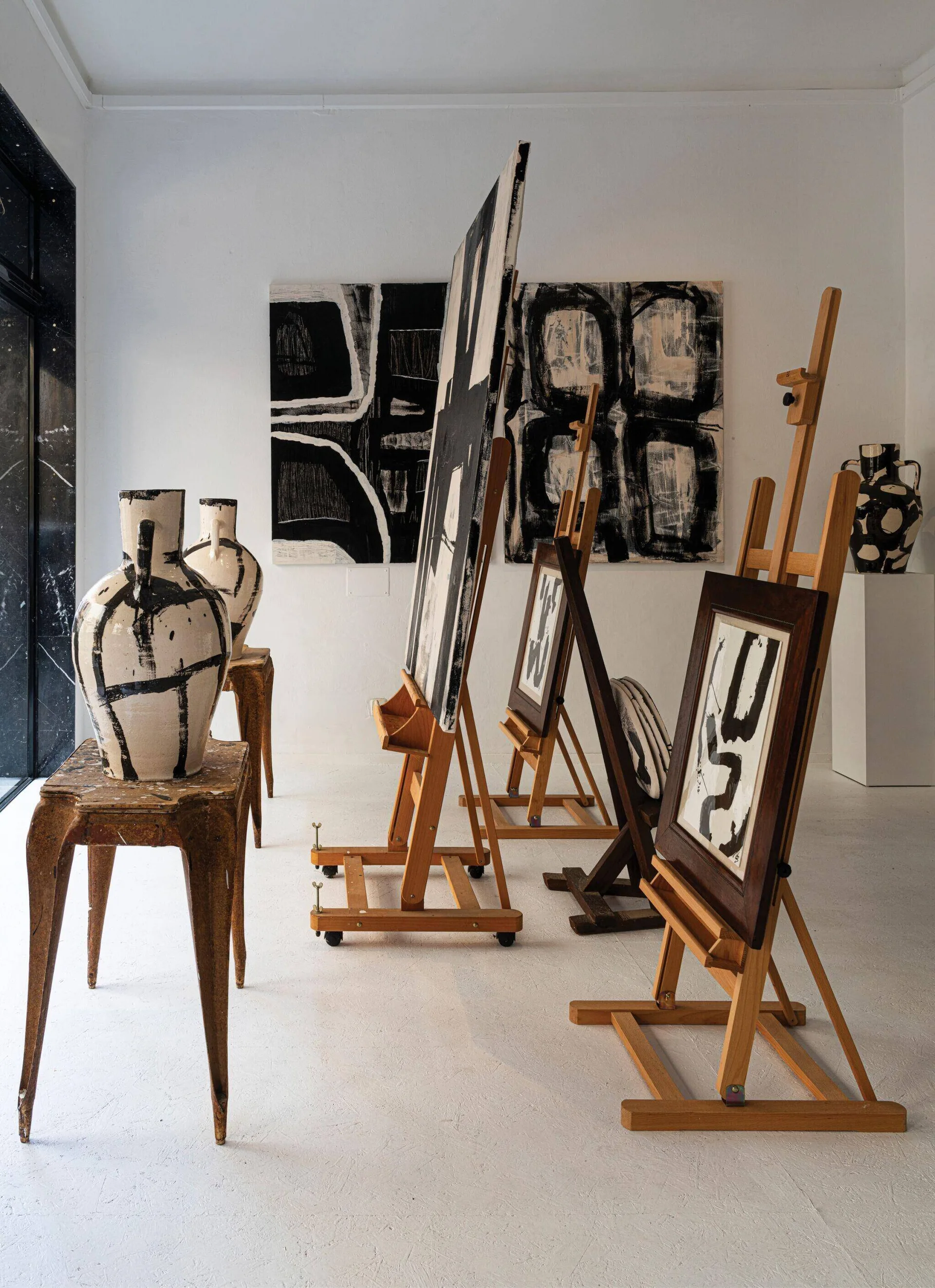 Wooden art easels displaying abstract black and white paintings stand on a white floor alongside patterned ceramic vases on distressed stools.