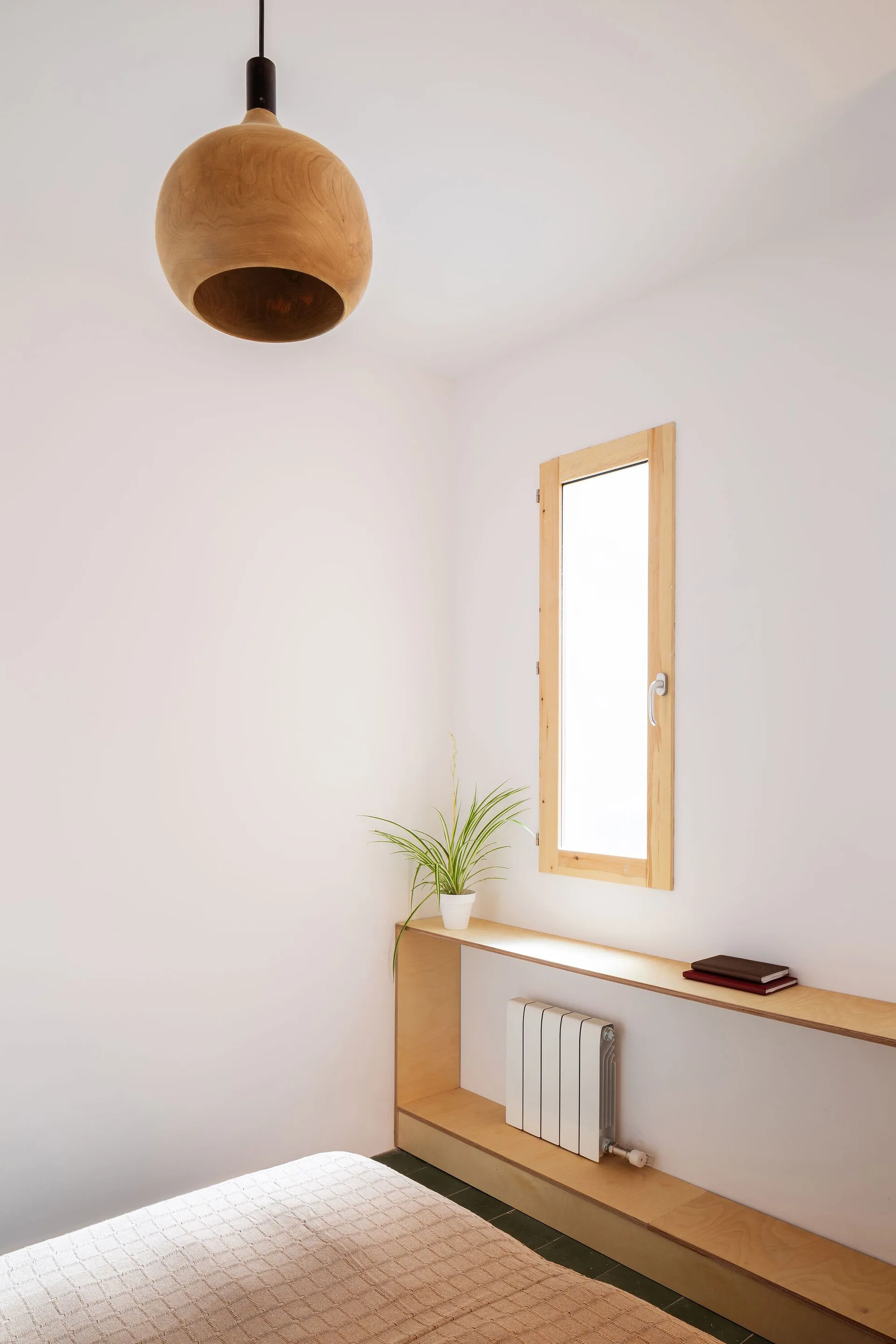 A minimalist bedroom featuring a wooden dome pendant light hanging above a custom plywood shelf built around a white radiator below a narrow window.
