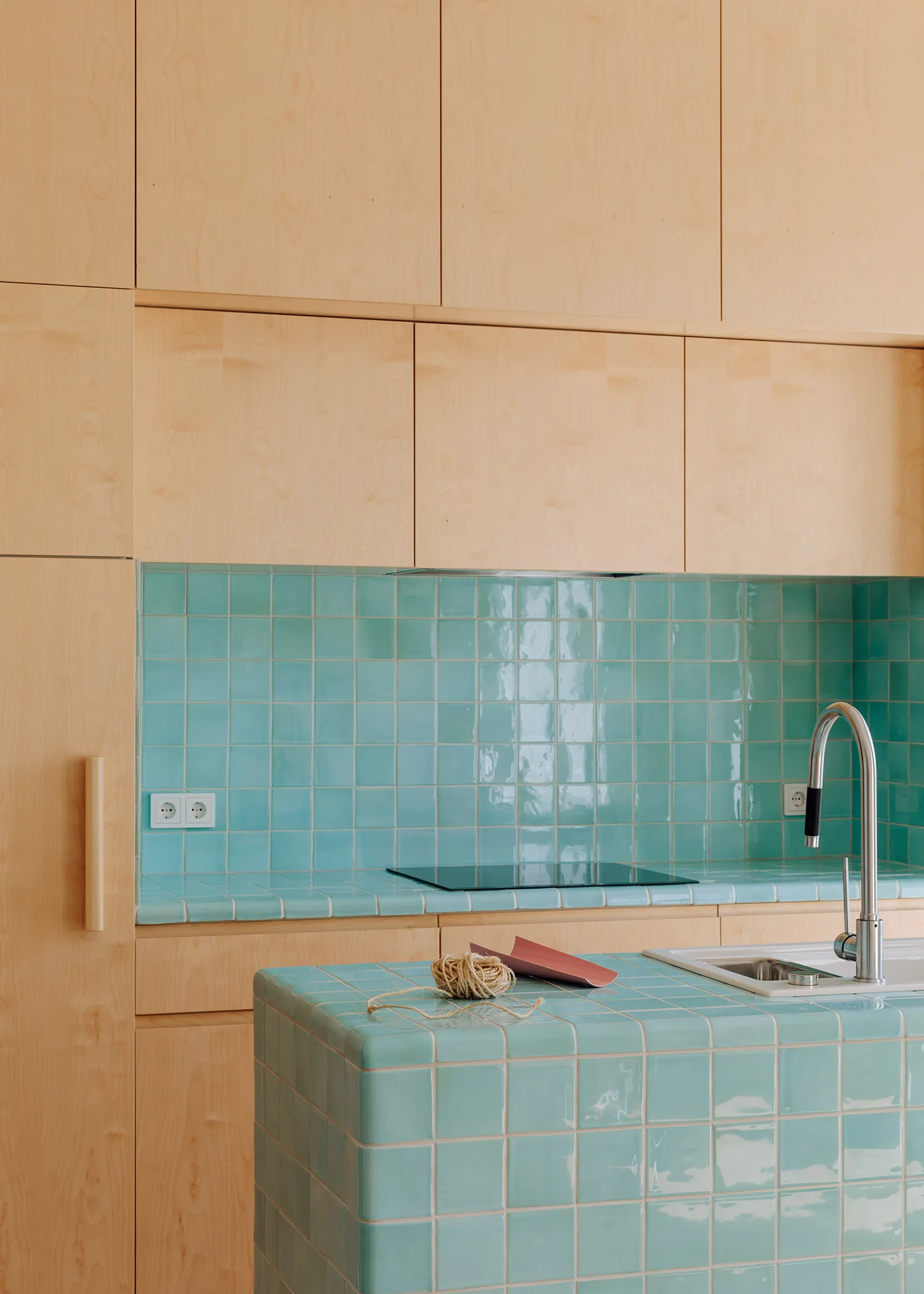 A modern kitchen featuring flat-front light wood cabinets, a vibrant turquoise tiled backsplash, and a matching tiled island with a silver faucet.