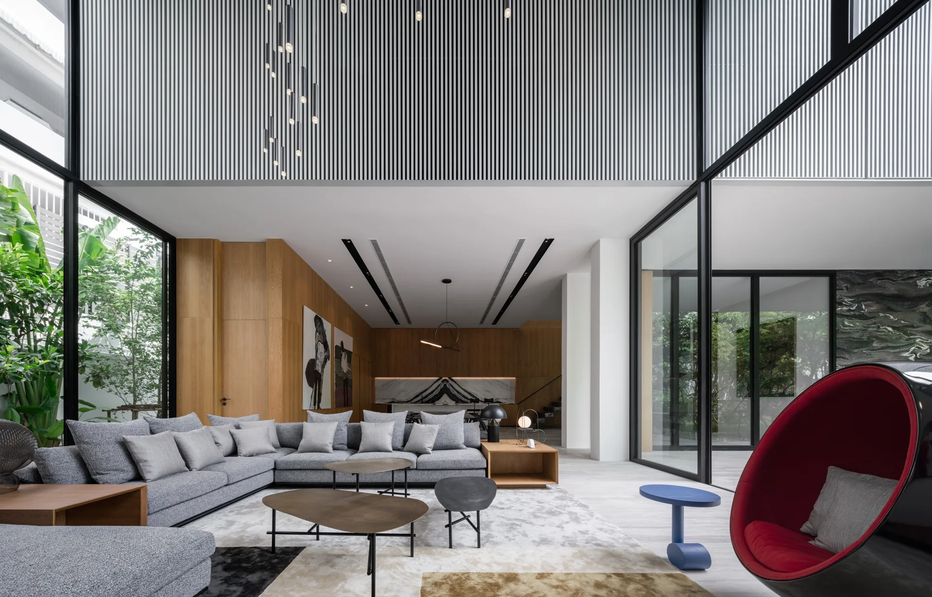 A spacious modern living room features a grey sectional sofa, tiered wood coffee tables, wood-paneled walls, and a black-and-red egg-shaped accent chair.