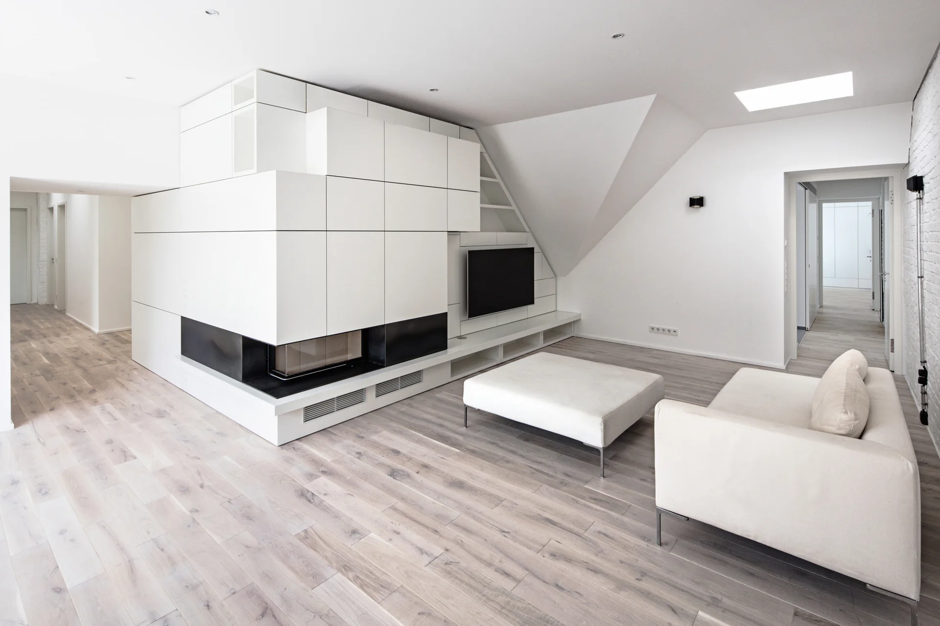 A minimalist living room featuring a large white geometric storage unit with an integrated fireplace, a light wood floor, and a white sofa.