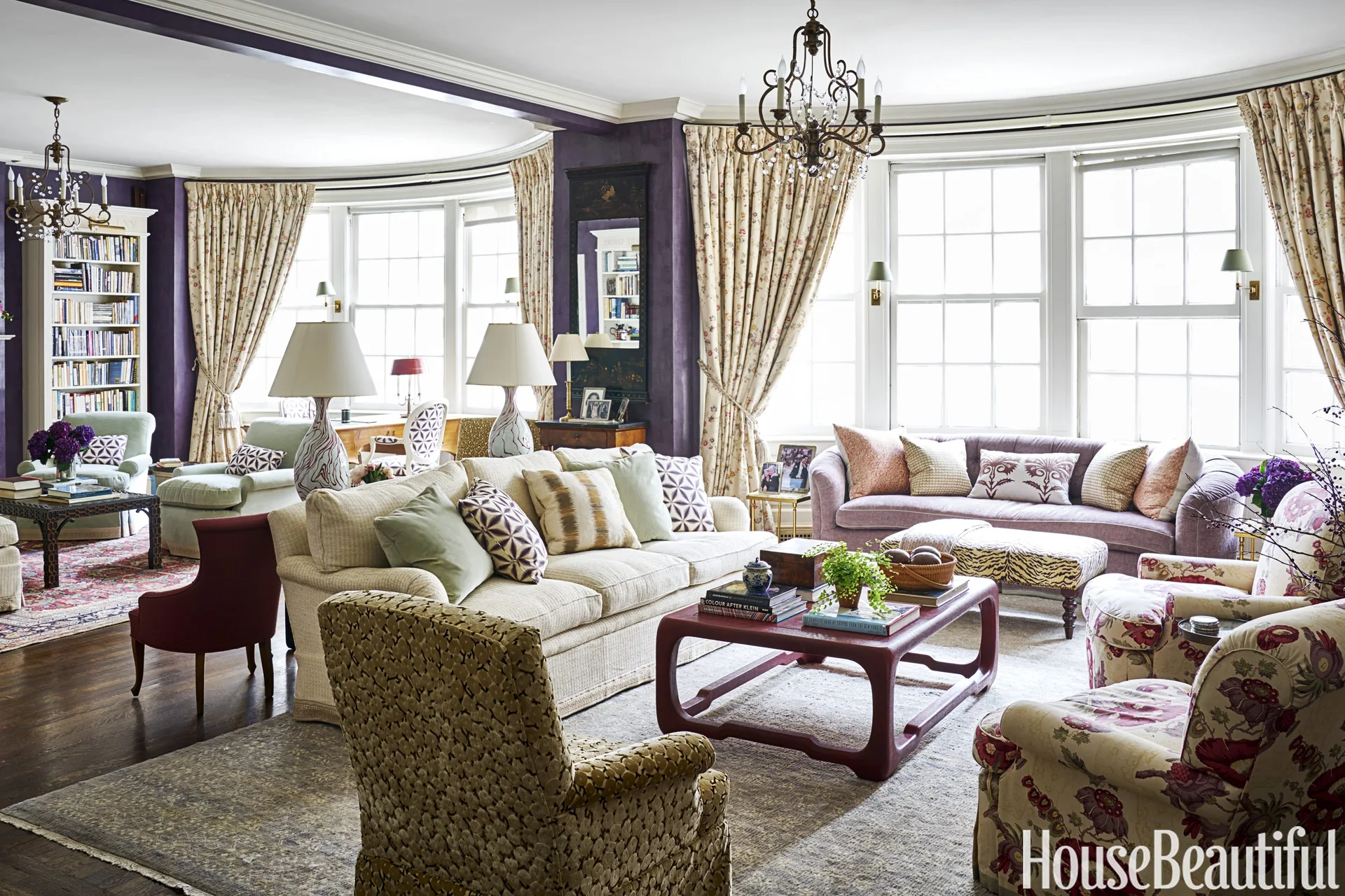 A maximalist living room featuring deep purple walls, a curved bay window with patterned drapes, multiple upholstered sofas, and a low red coffee table.