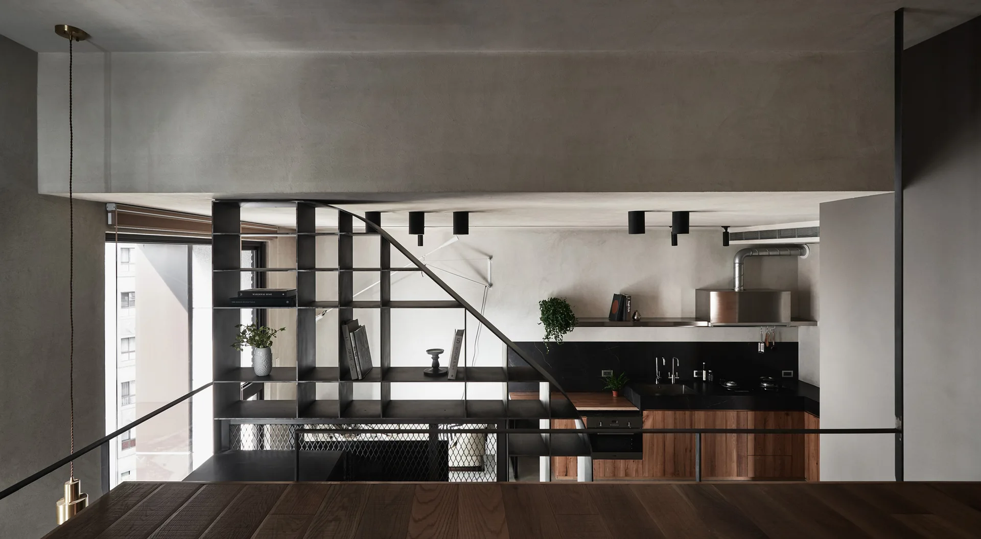 A modern industrial kitchen features wood cabinets and a black countertop viewed behind a geometric black metal shelving unit and grey plaster walls.