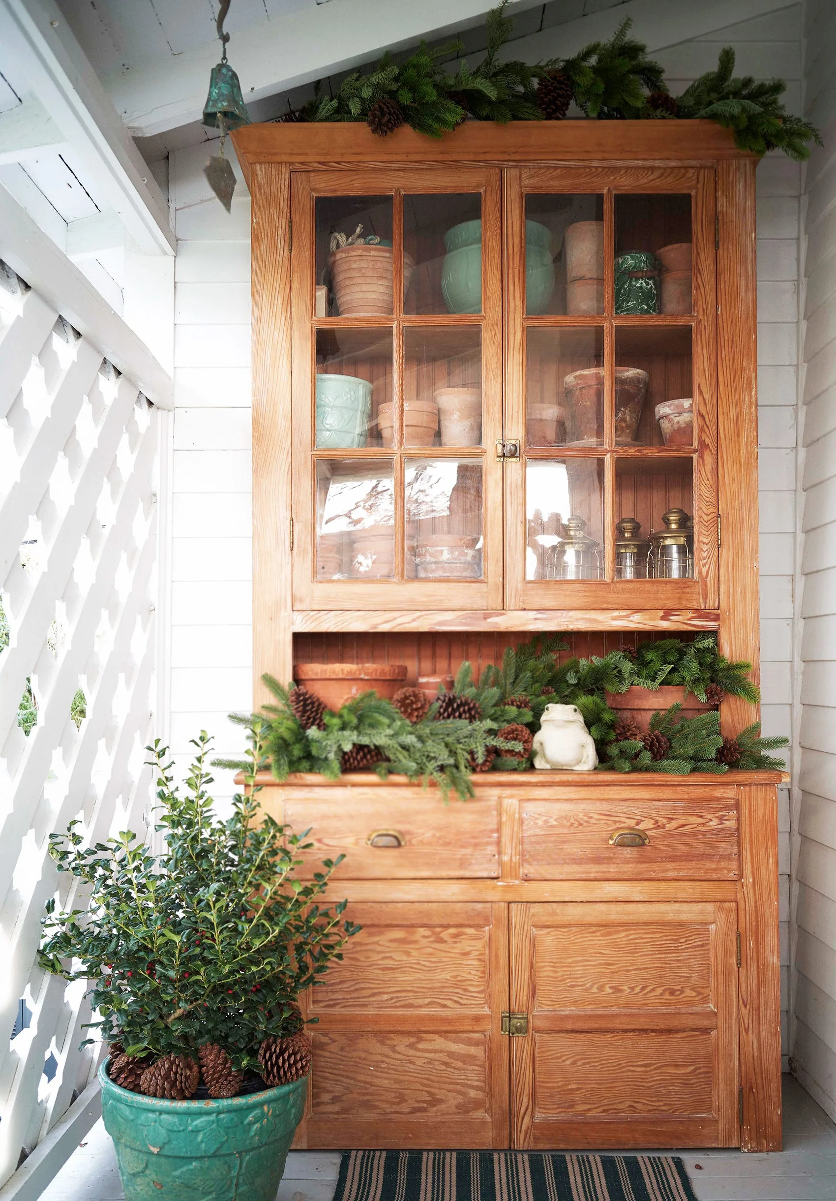 A rustic wooden hutch with glass doors is decorated with evergreen pine garlands and sits on a white porch with horizontal siding.