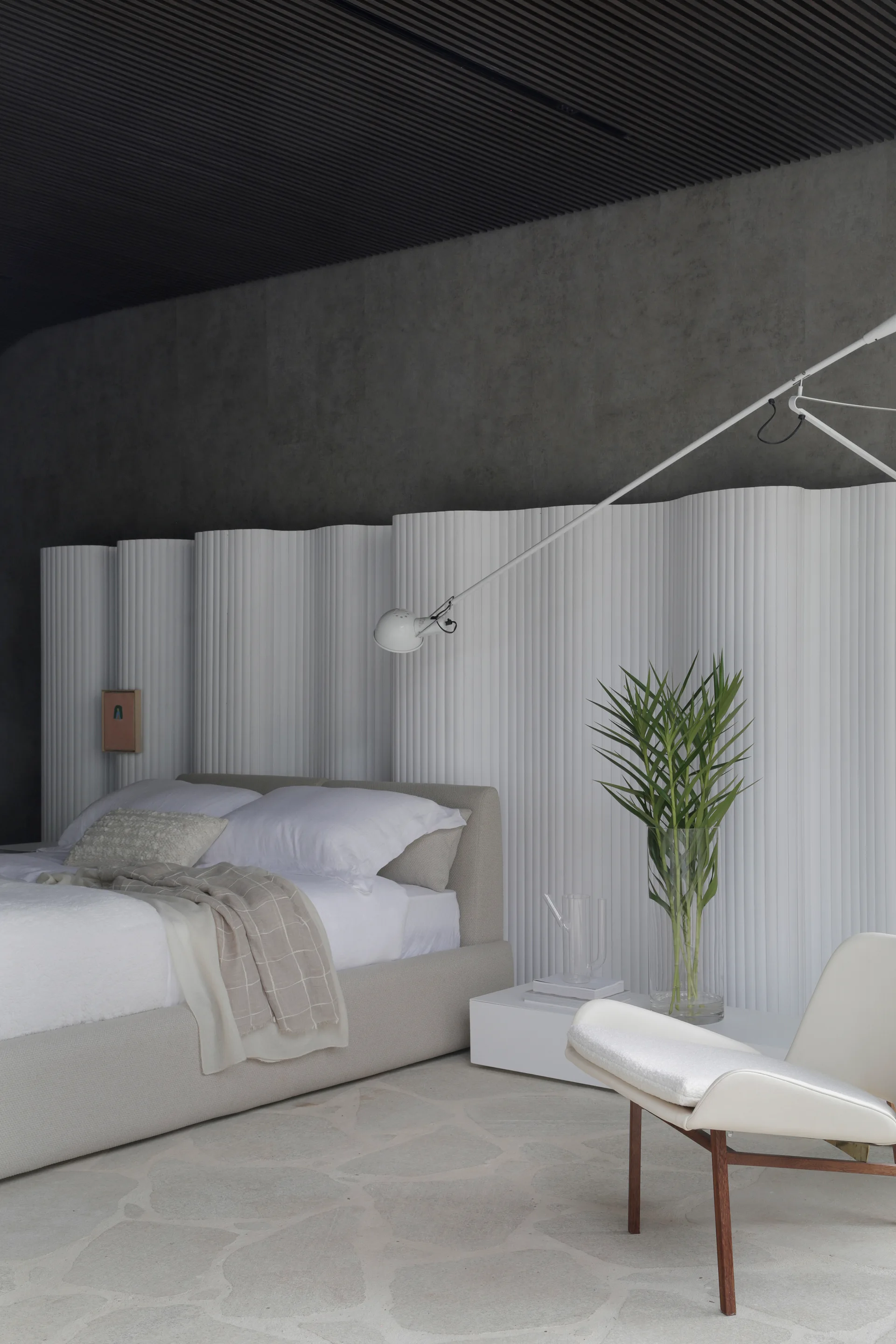 A minimalist bedroom featuring a curved white fluted wall panel behind an upholstered bed, a dark slatted ceiling, and irregular stone floor tiles.