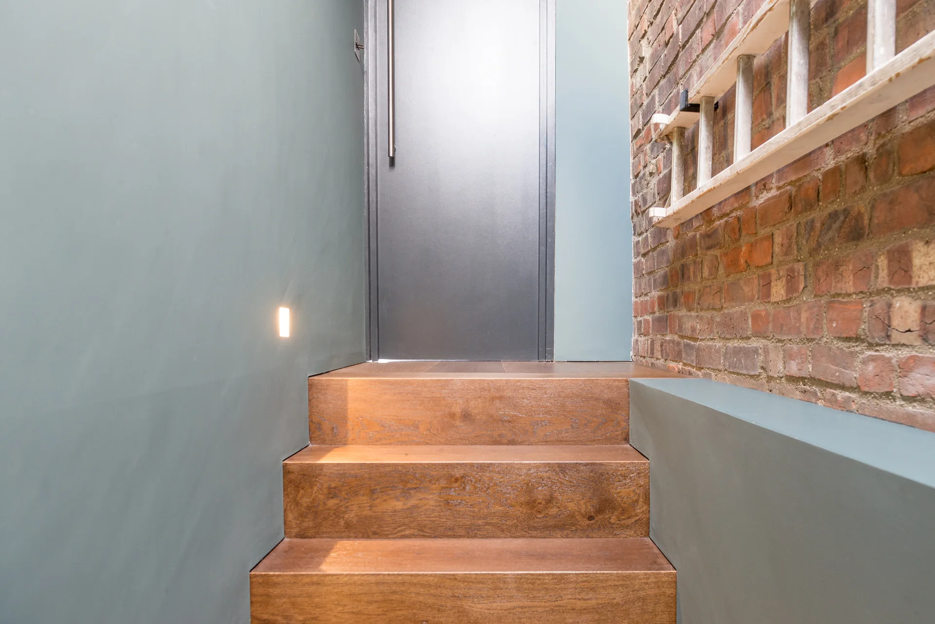 Wooden stairs lead up to a dark gray door flanked by a smooth green-gray wall with a step light and an exposed brick wall with a ladder.
